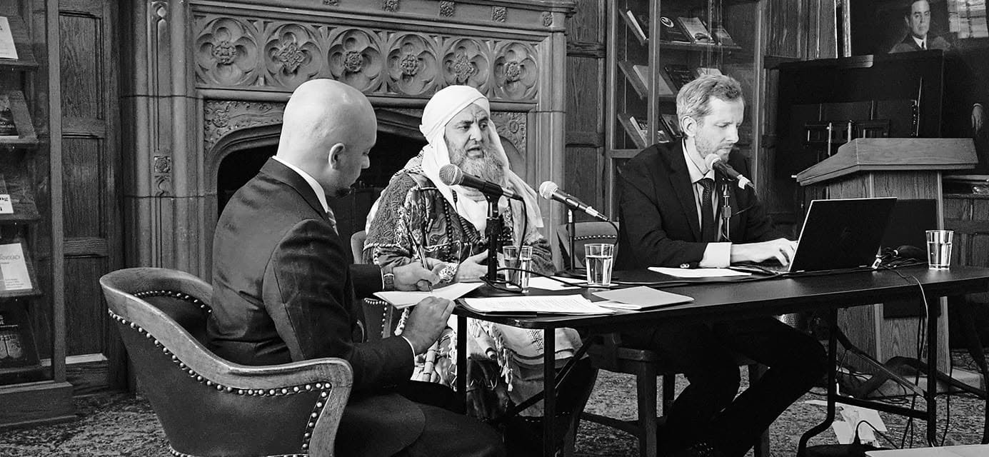 Three men sit at a table in a wood-paneled room with an ornate decorated wood wall behind them. The middle man, dressed in traditional Middle Eastern attire, is speaking into two microphones. The man on the left is taking notes, and the man on the ri