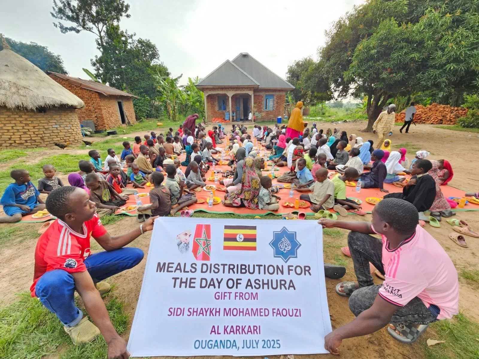 A large outdoor gathering of children and adults sitting on mats, eating meals, during a community event in Uganda. Two people hold a sign indicating the event is a food distribution for Ashura, organized by Sidi Shaykh Mohamed Faouzi Al Karkari in J