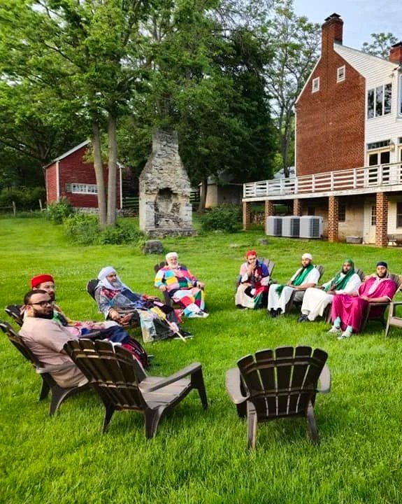 People dressed in colorful traditional clothing, sitting in a semi-circle on the grass in a backyard with trees, a brick house, and a stone fireplace.