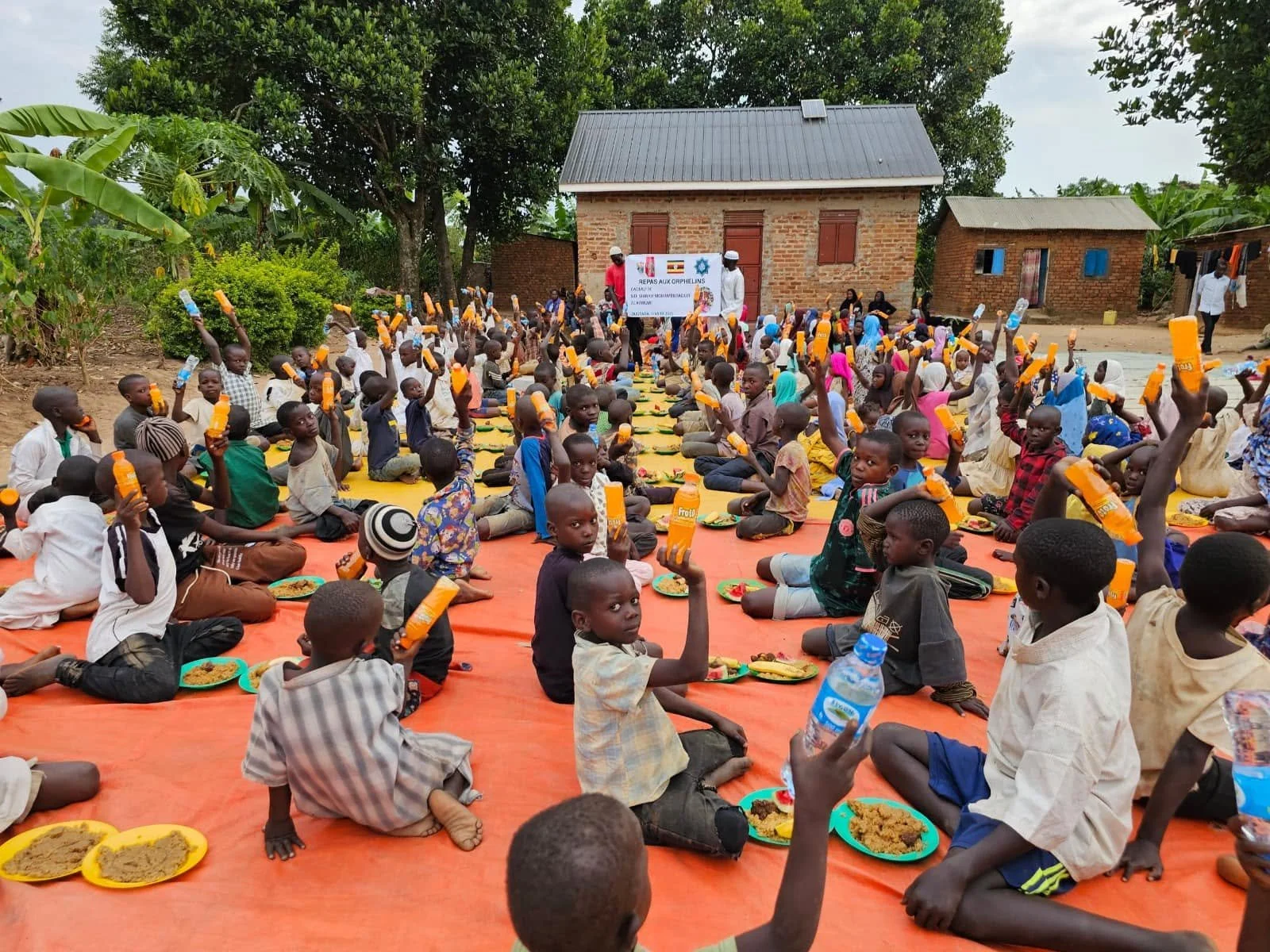 Children sitting on colored mats outdoors, raising bottles of orange-colored drink during a communal meal or gathering, with a house and trees in the background.