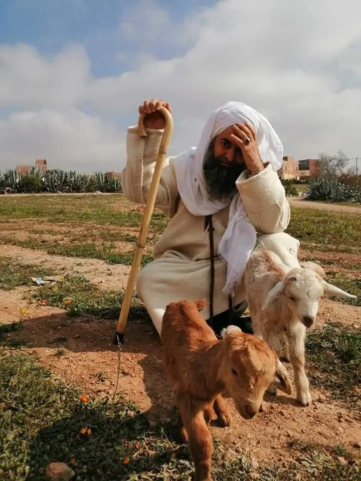 A man dressed in traditional Middle Eastern attire, sitting on the ground in a desert landscape with goats, holding a shepherd's staff, with his hand on his forehead.