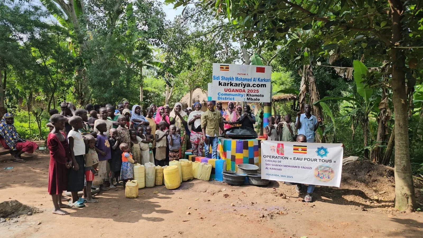 Community gathering of children and adults in a rural outdoor setting with water donation supplies, a sign promoting a water project, and a banner for a charity operation in Uganda.