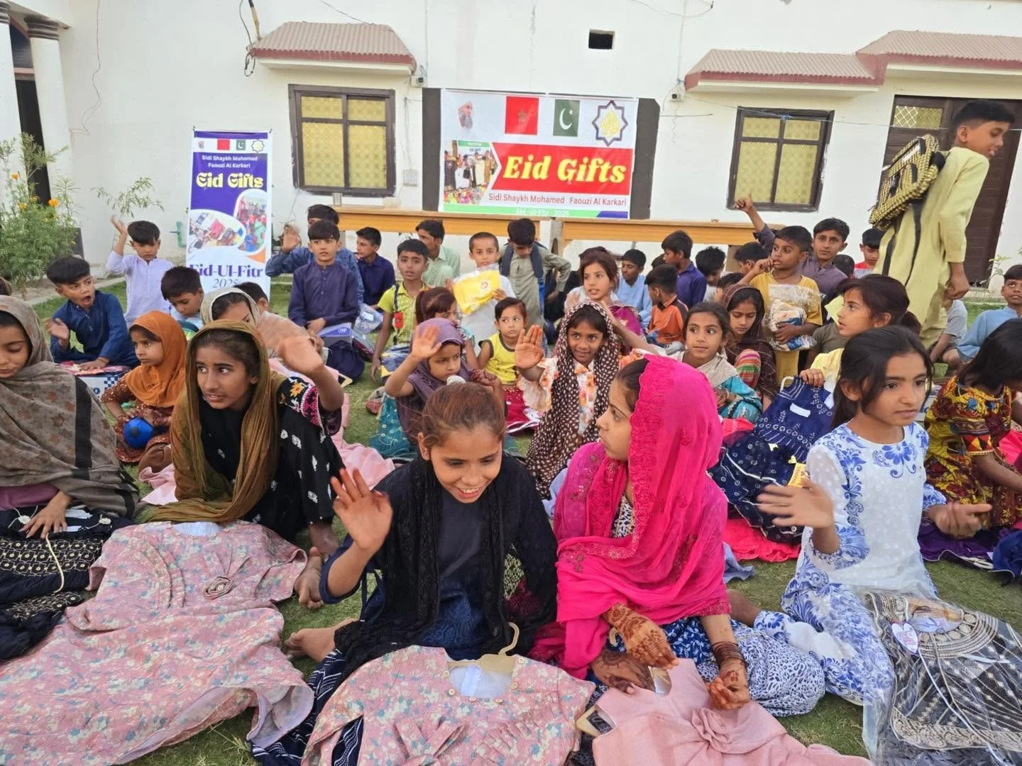 Group of children and women gathered outdoors for Eid celebration, sitting on the grass, some waving and holding gifts, with banners and buildings in the background.