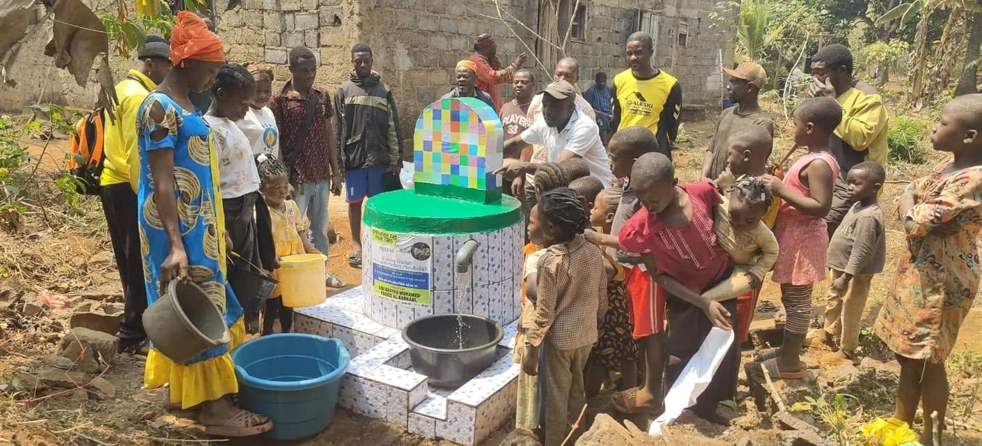 Group of people, including children, gathered around a newly constructed water well with a tiled surface and a colorful roof, in a rural outdoor setting.
