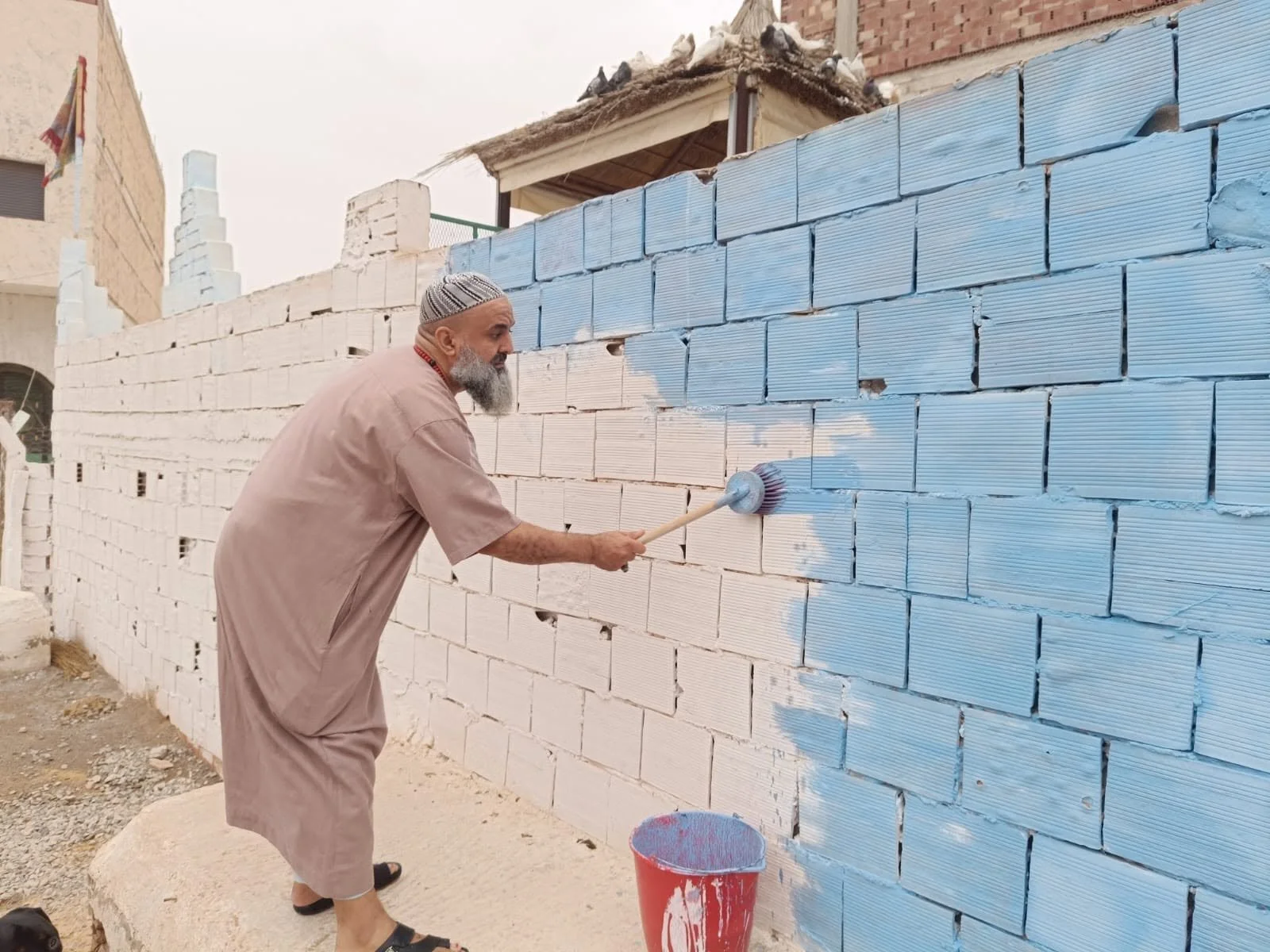 A man wearing traditional Middle Eastern clothing painting a brick wall with blue paint, standing on a concrete step using a roller brush, with a red bucket of paint nearby.