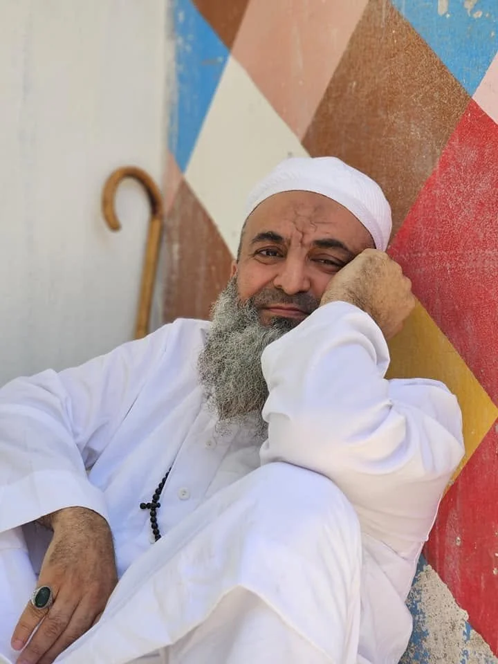 An elderly man with a long gray beard, wearing a white shirt and white cap, sitting against a colorful, geometric wall, with a wooden cane in the background.