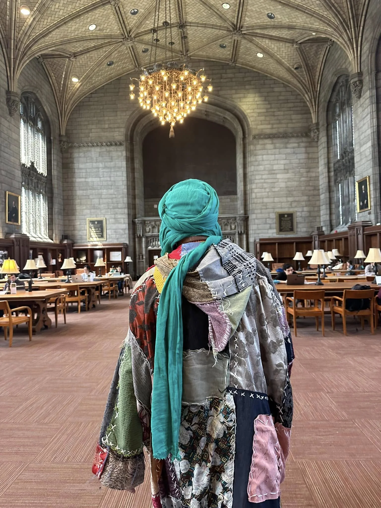A person with a teal head covering and a multicolored patchwork quilted coat standing inside a grand historic library with high vaulted ceilings, large chandelier, tall windows, and wooden bookshelves with lamps.