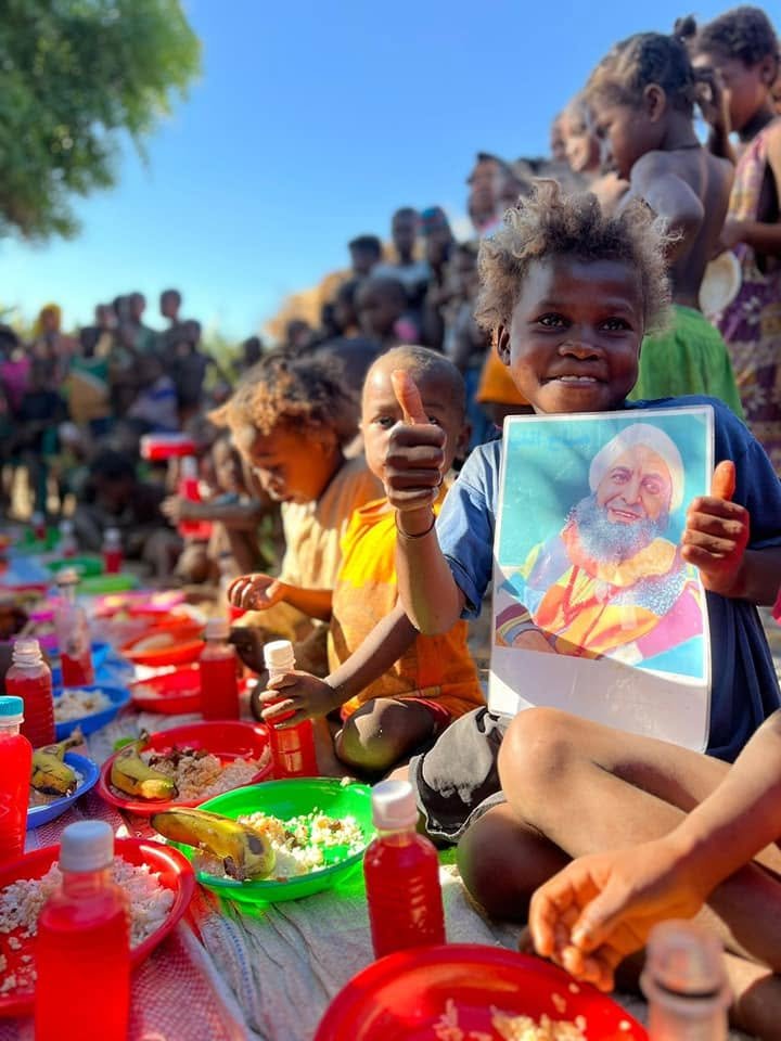 A group of children sitting on a cloth with plates of food and bottles of drink, outdoors under a bright blue sky. One child is holding a picture of a guru and giving a thumbs-up.
