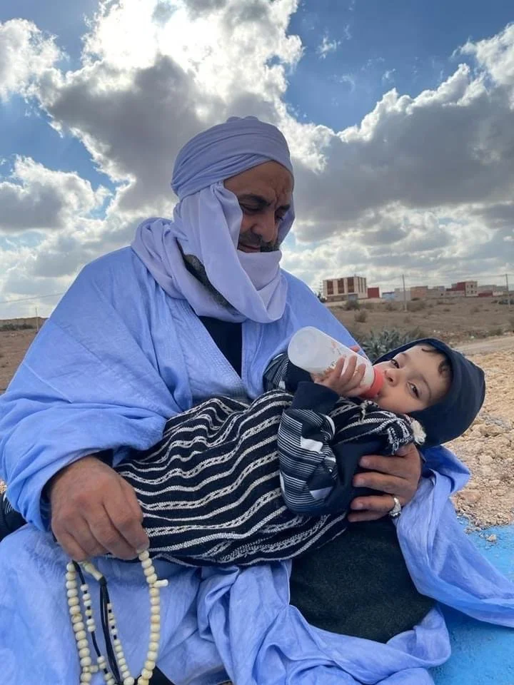 Man is holding and feeding a young boy with a bottle outside during daytime, in a desert-like area with buildings and clouds in the sky.