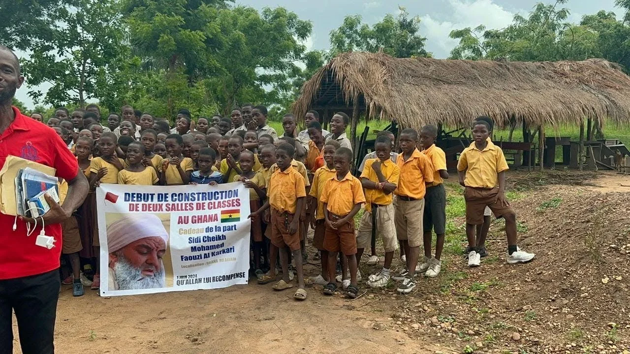 Group of children in yellow and brown school uniforms gathered outdoors, holding a banner in front of a thatched-roof structure, with trees in the background.