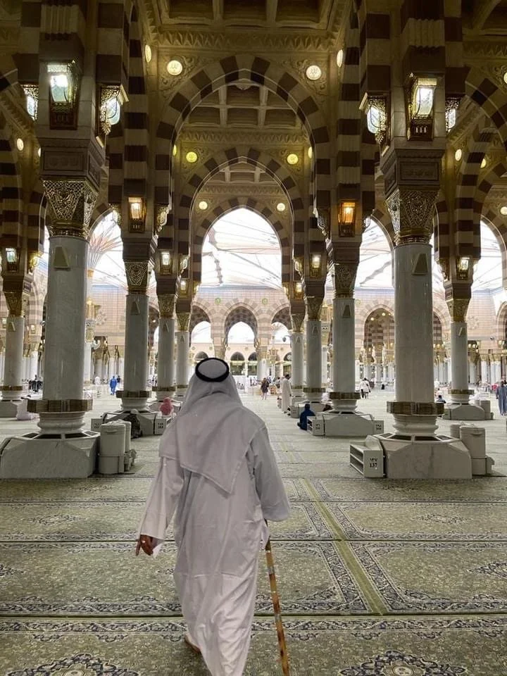 A man dressed in traditional Middle Eastern attire walking inside a grand mosque with intricate architecture, large arches, and ornate hanging lamps.