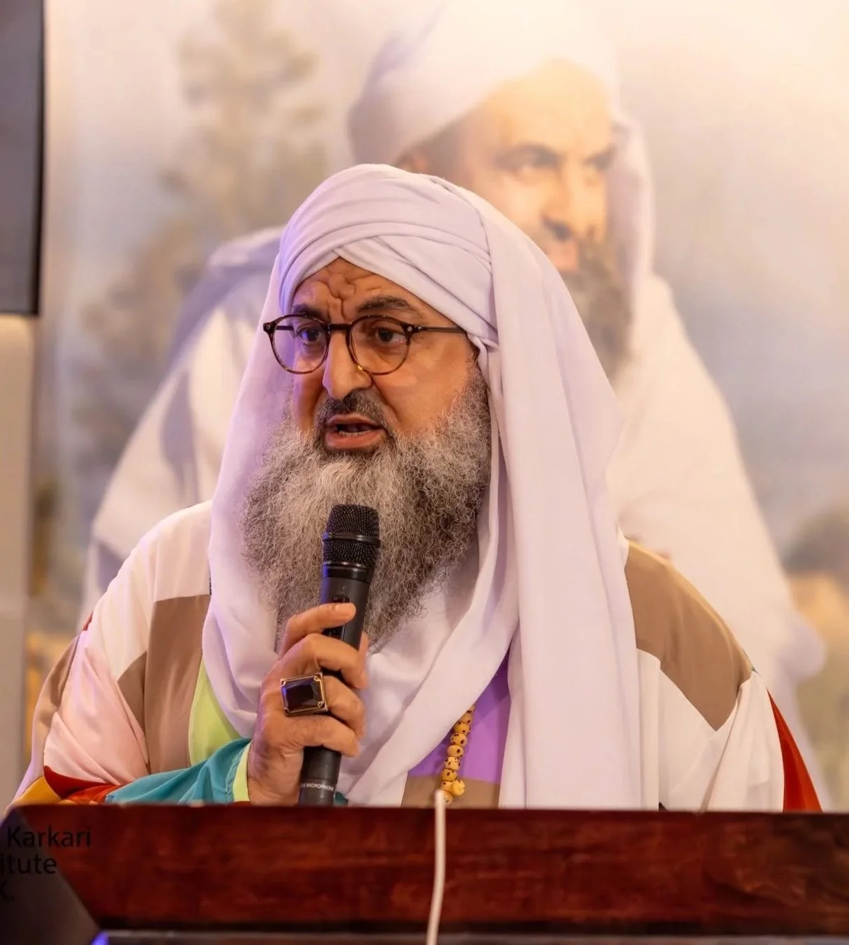 A man with glasses and a long gray beard, dressed in traditional attire with a white turban, speaking into a microphone. A large portrait of another man, also dressed in traditional attire with a white turban, is visible in the background.