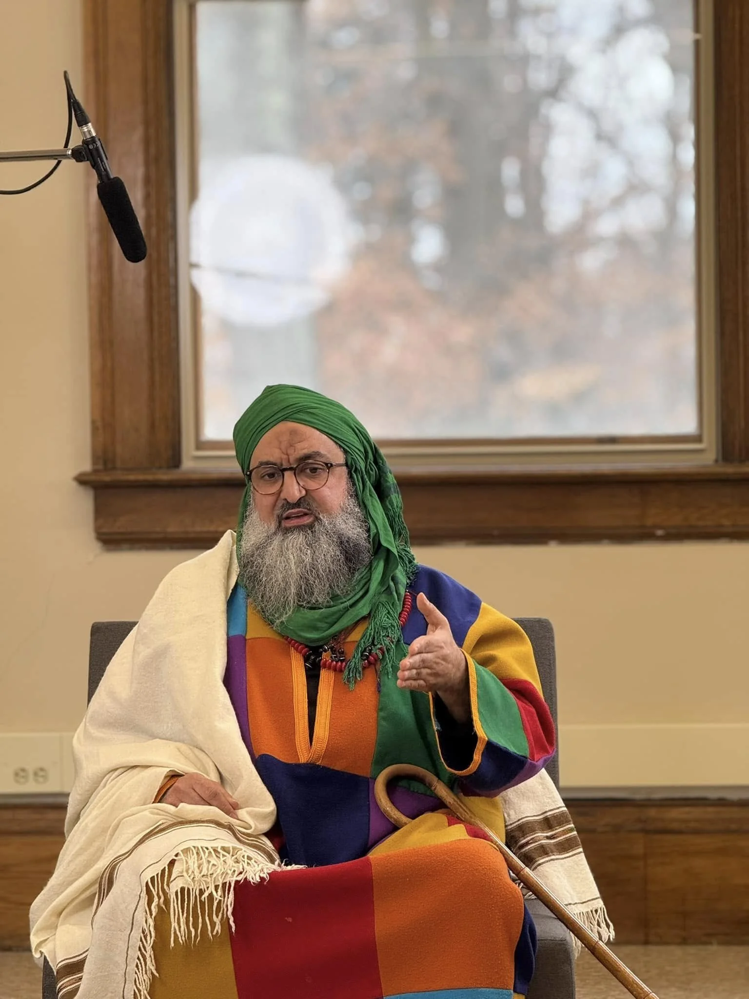 A man with glasses and a long gray beard wearing a colorful jacket, green headscarf, and a beige shawl sitting in a room with a window. He is gesturing with his right hand and sitting in front of a plain wall with a window showing trees outside.