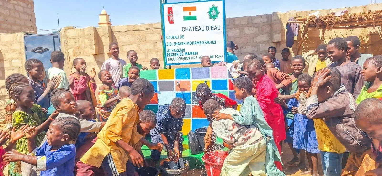 Group of children gathered around a colorful water fountain, playing and splashing water on each other in an outdoor setting with brick walls and a sign in French in the background.
