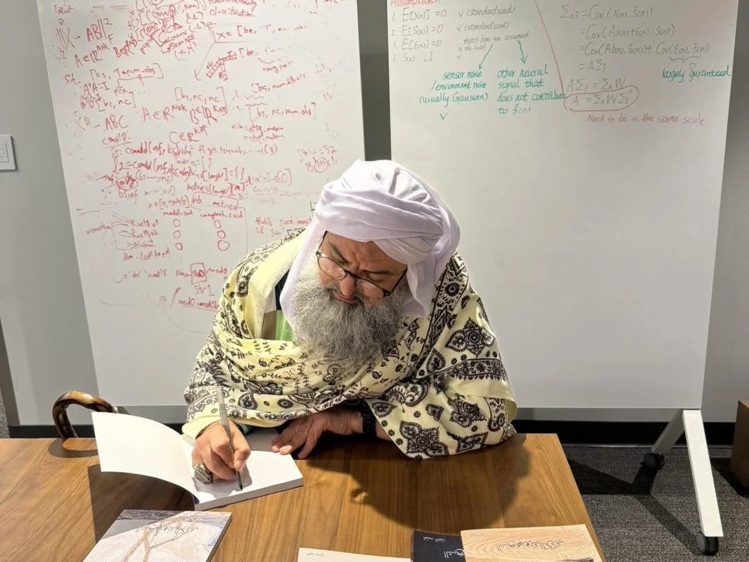 An elderly man with a white turban, glasses, and a patterned shawl is sitting at a wooden table, writing in a notebook with a pen. Behind him is a whiteboard filled with handwritten notes and diagrams in red, green, and black ink.