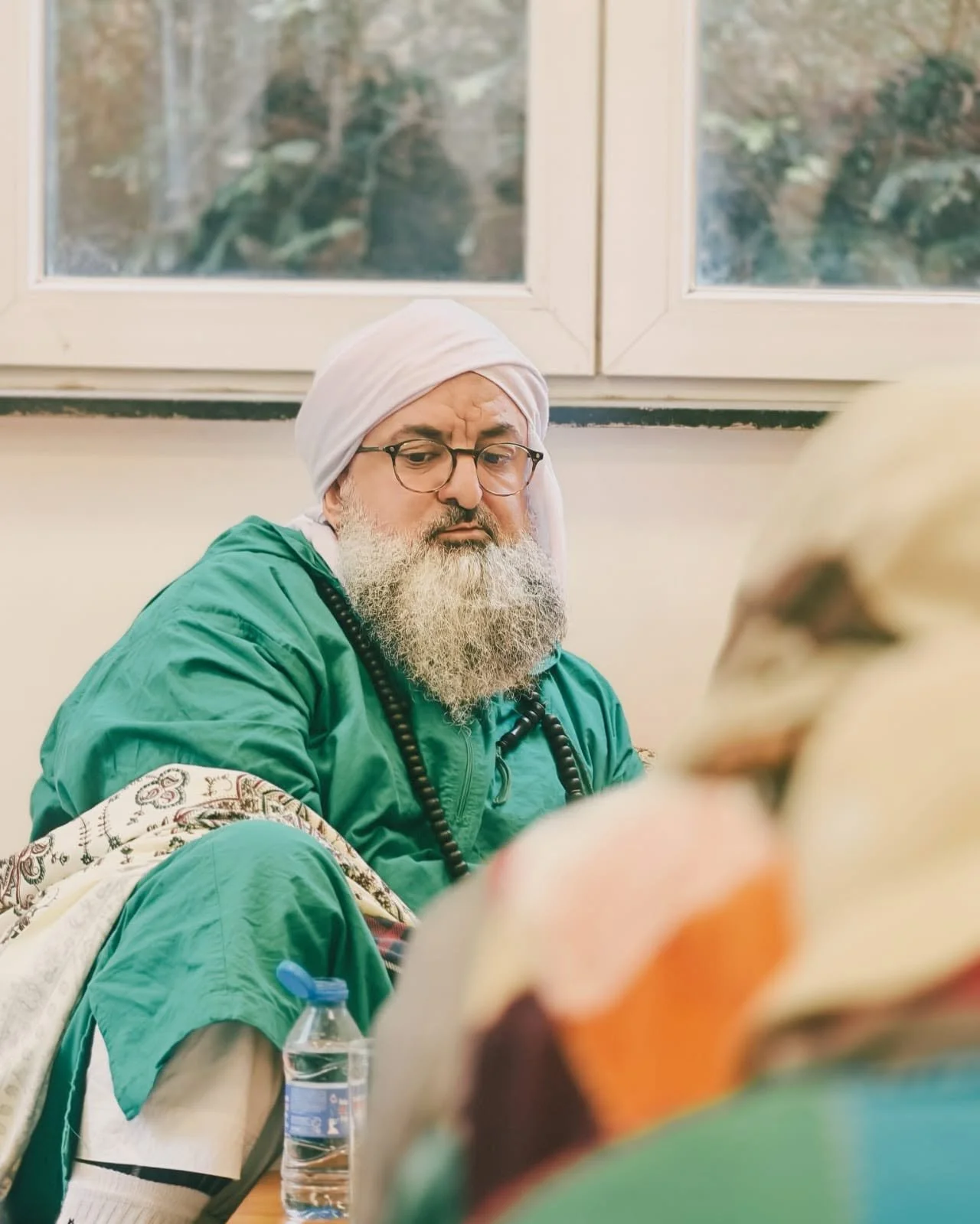 A man with a white beard, glasses, and a white turban, wearing a green jacket, sitting indoors near a window, looking down thoughtfully.