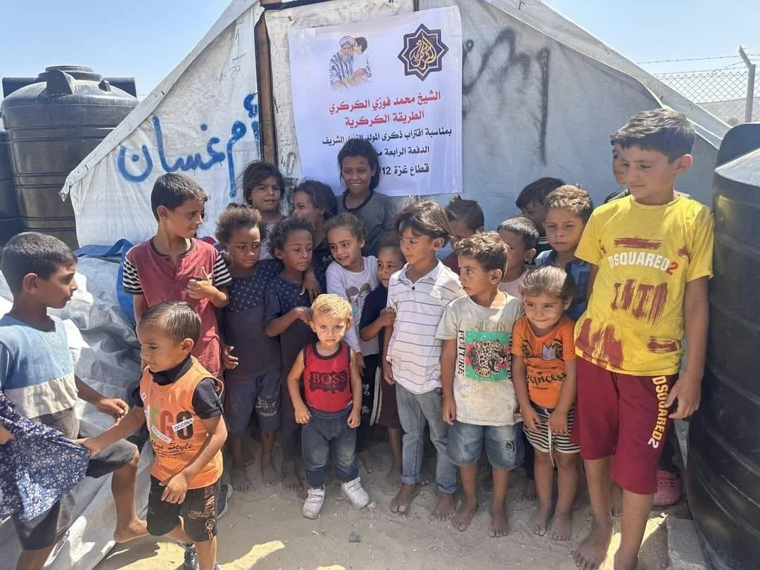 Group of children standing in front of a tent and a water tank, with a sign containing Arabic text and a logo, in a desert or arid environment.