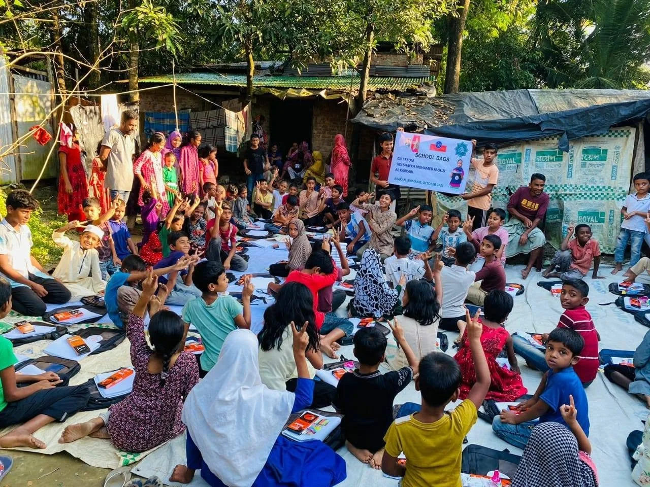 A large group of children sitting on a white sheet outdoors, many raising their hands, with teachers and adults standing around them. Some children have school bags and notebooks. There is a signboard indicating a school-related event, and makeshift 