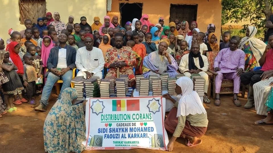 A large group of people gathered outdoors, with some seated in front and others standing behind. In front are several stacks of books and two women arranging or presenting the books. There is a sign with the flags of Cameroon and China, and text in F