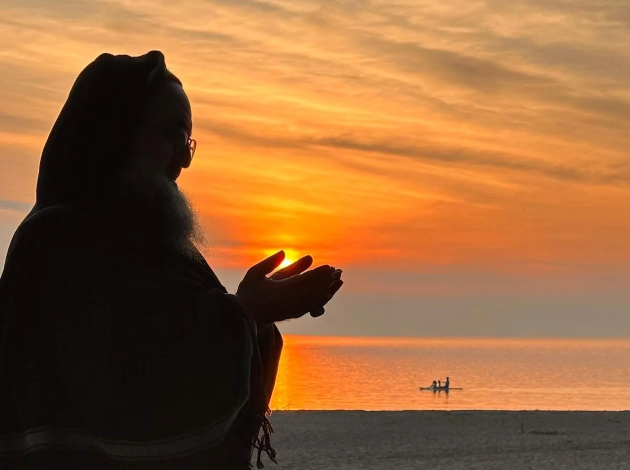 A silhouette of a man holding his hands together in a prayer gesture during sunset at the beach, with a small boat on the water in the background.