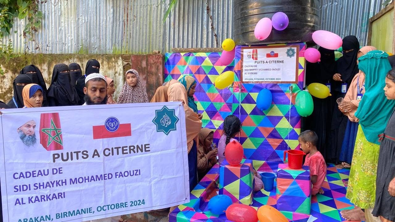 Group of people, including children and women, gather around a colorful display with balloons, boxes, and a sign in Arabic and French. A large banner with Moroccan, Mauritanian, and Islamic symbols is present.