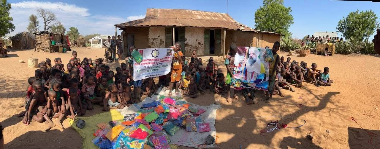 Children gathered outdoors in front of a modest house in a rural area, receiving supplies and aid, with banners and a large portrait of a man on display.