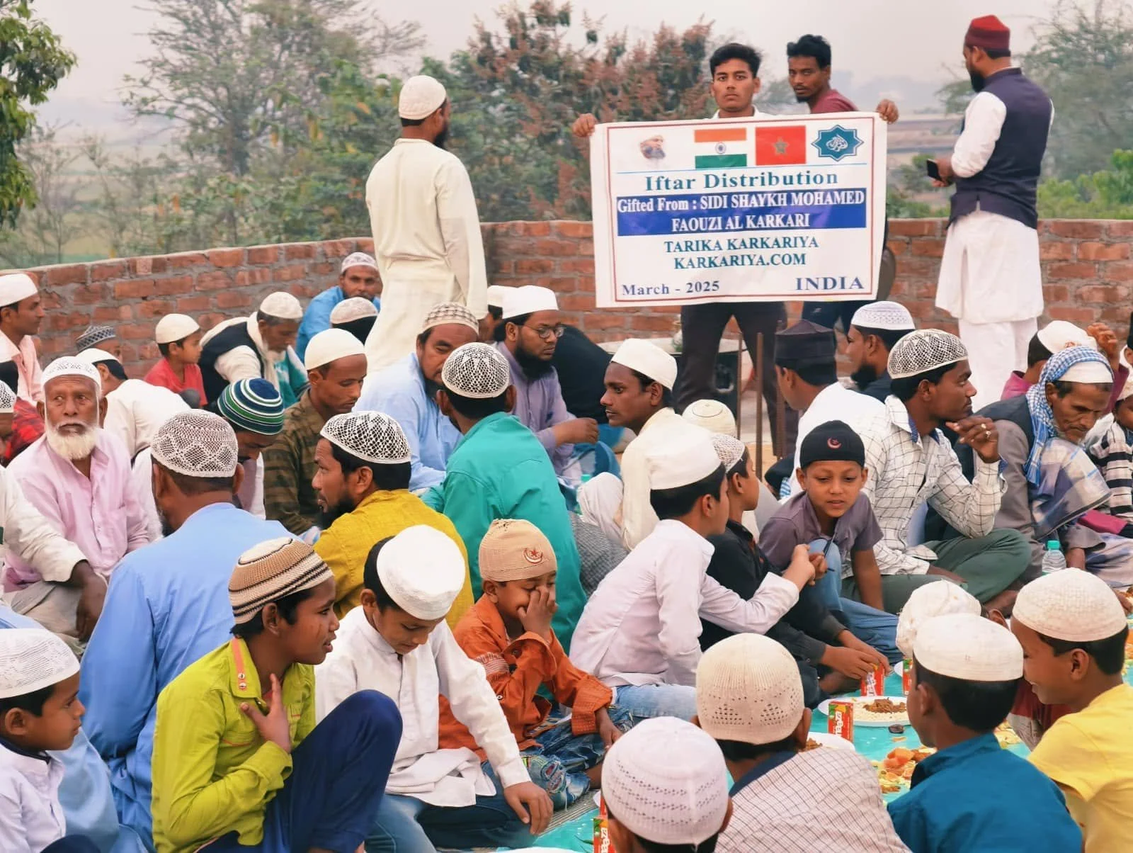 A large group of men and boys seated on the ground, wearing traditional Muslim attire including various types of caps, during an outdoor event in India. A few men are standing, holding a sign that reads 'Iftar Distribution Gifted from Sidi Shaykh Moh