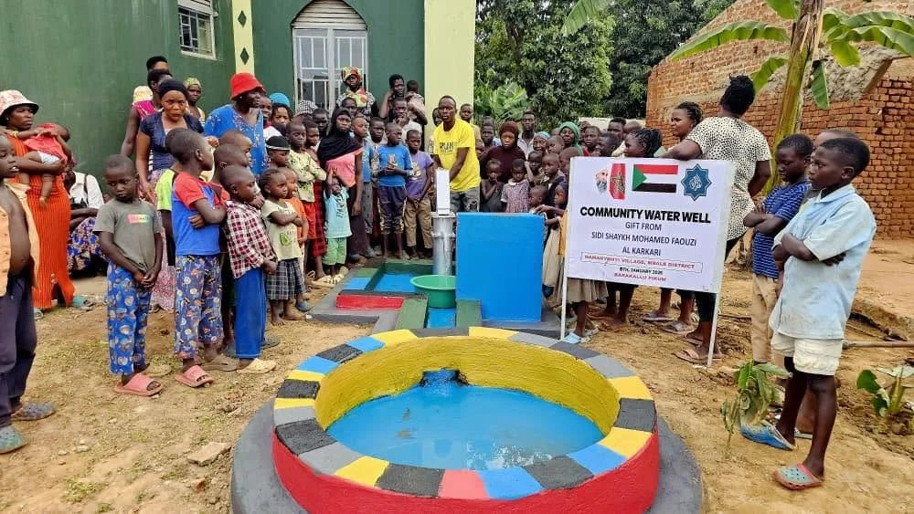 A community gathering around a newly installed water well in an outdoor setting, with children and adults present, holding a sign indicating donation from Sid Shaykh Mohamed Fauzi Al Karkari.