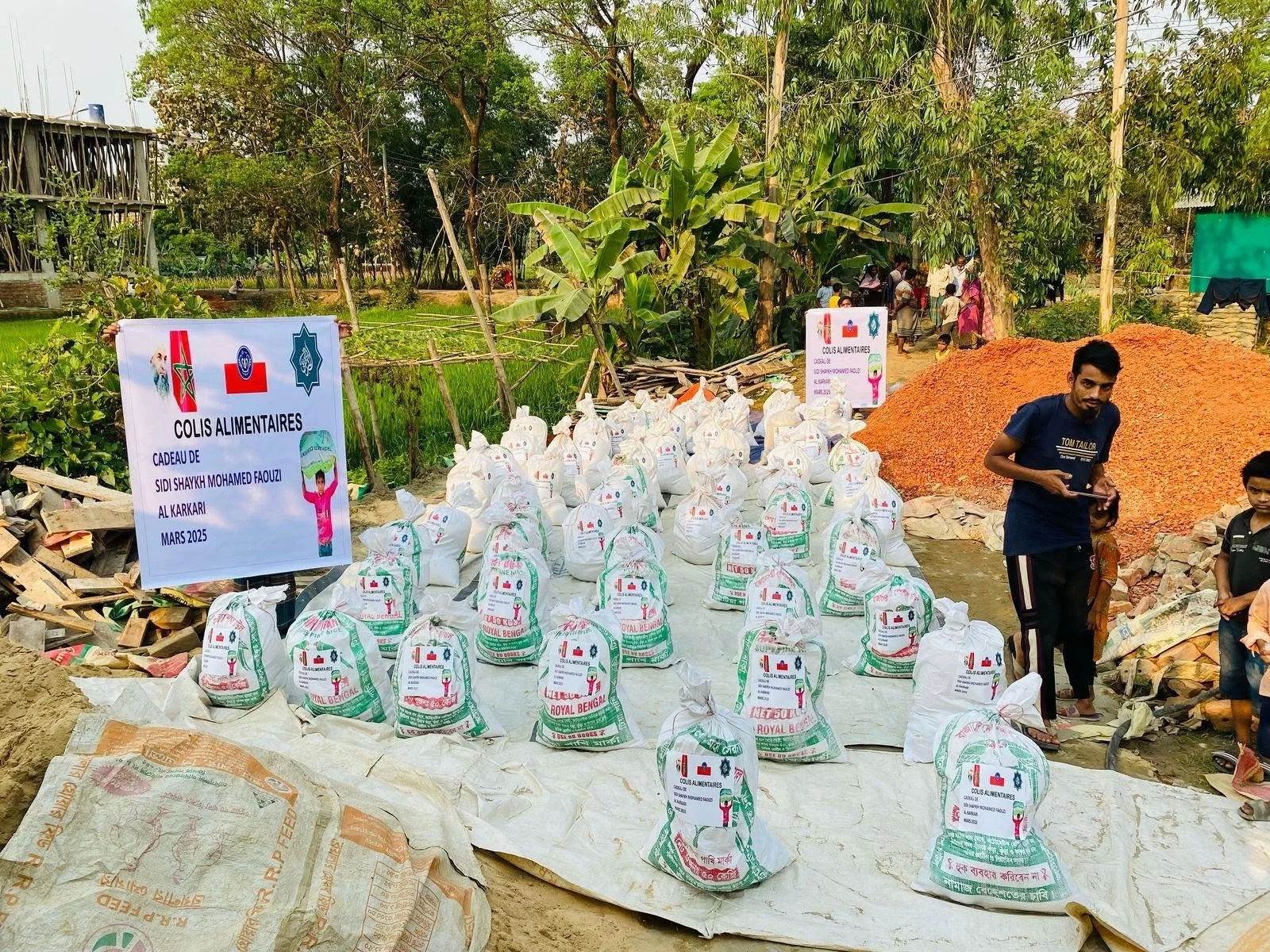 Community food aid distribution with bags of supplies arranged in rows, a sign indicating it is part of a project from March 2025, and people standing around in a rural setting with trees and construction materials nearby.