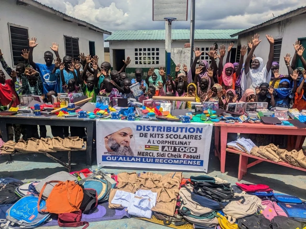 Group of children and women standing behind tables filled with school supplies, smiling and raising their hands in a welcoming gesture, in an outdoor schoolyard setting.