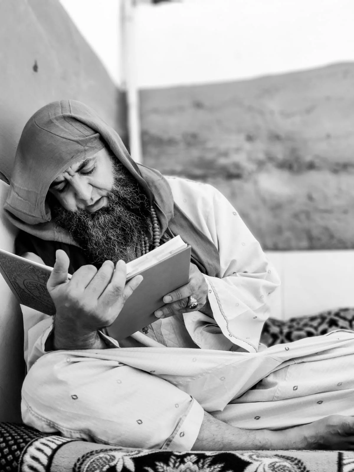 A man with a beard, wearing a hoodie and traditional attire, sitting and reading a book.