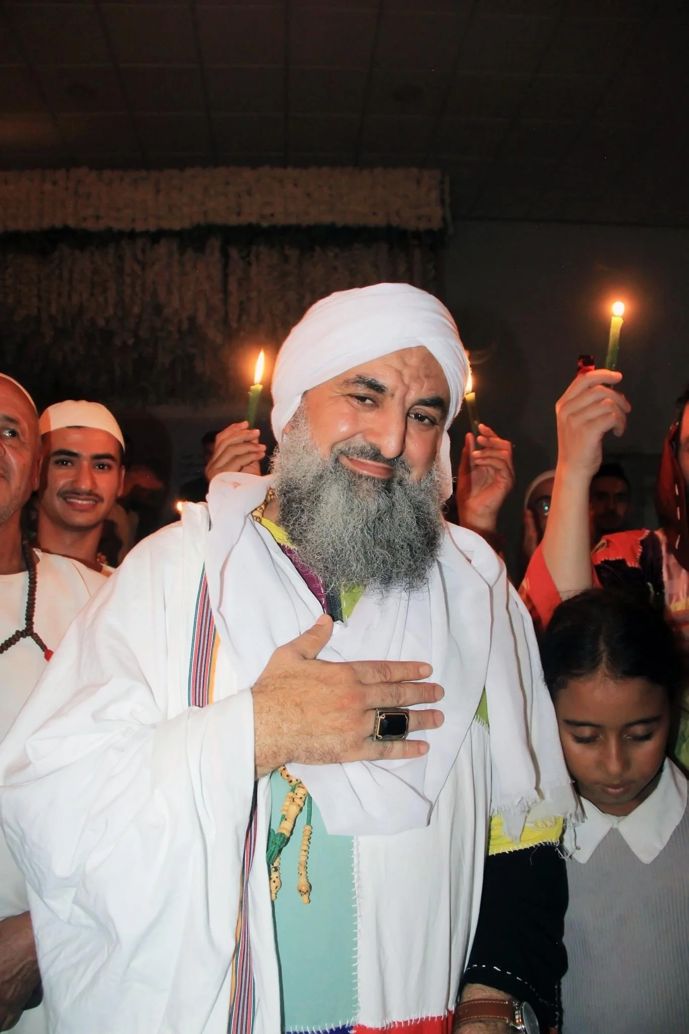 A group of people, mostly men, at a religious or cultural event. The central figure is an older man with a white beard, wearing a white turban and traditional white attire, holding his right hand over his chest. Others in the background are holding l
