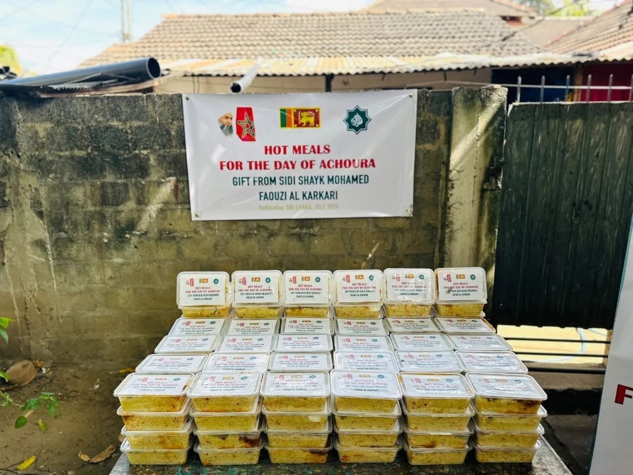 Plastic containers filled with hot meals stacked on a table, with a banner above reading "Hot Meals for the Day of Achoura" and mentioning a gift from Sidi Shayk Mohamed Faouzi Al Karkari, during a charity event in Emtatasha, Sri Lanka, July 2023.