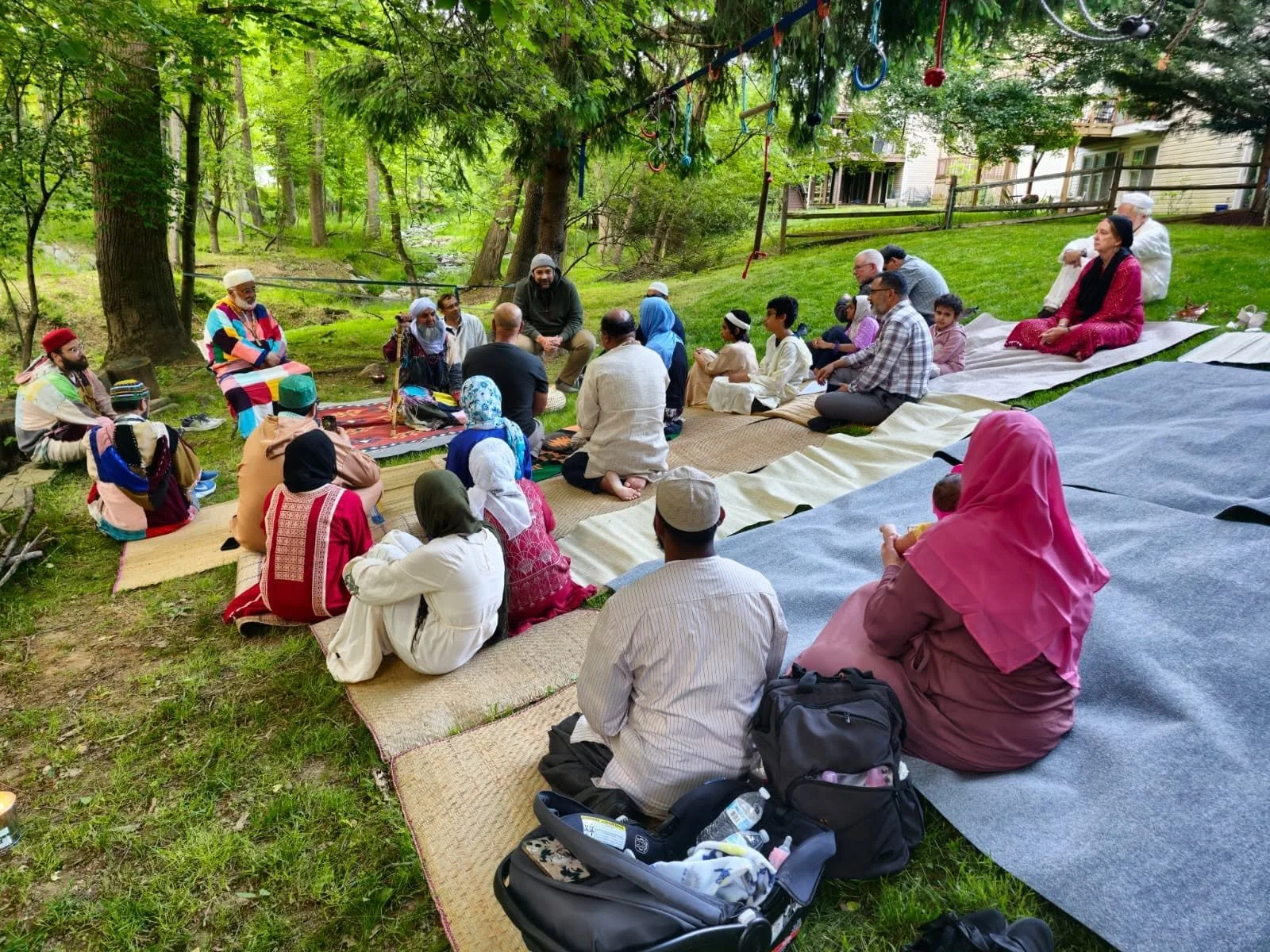 A diverse group of people sitting on mats and blankets outdoors in a wooded area, participating in a communal gathering or prayer session, with some wearing traditional clothing and others in casual attire.
