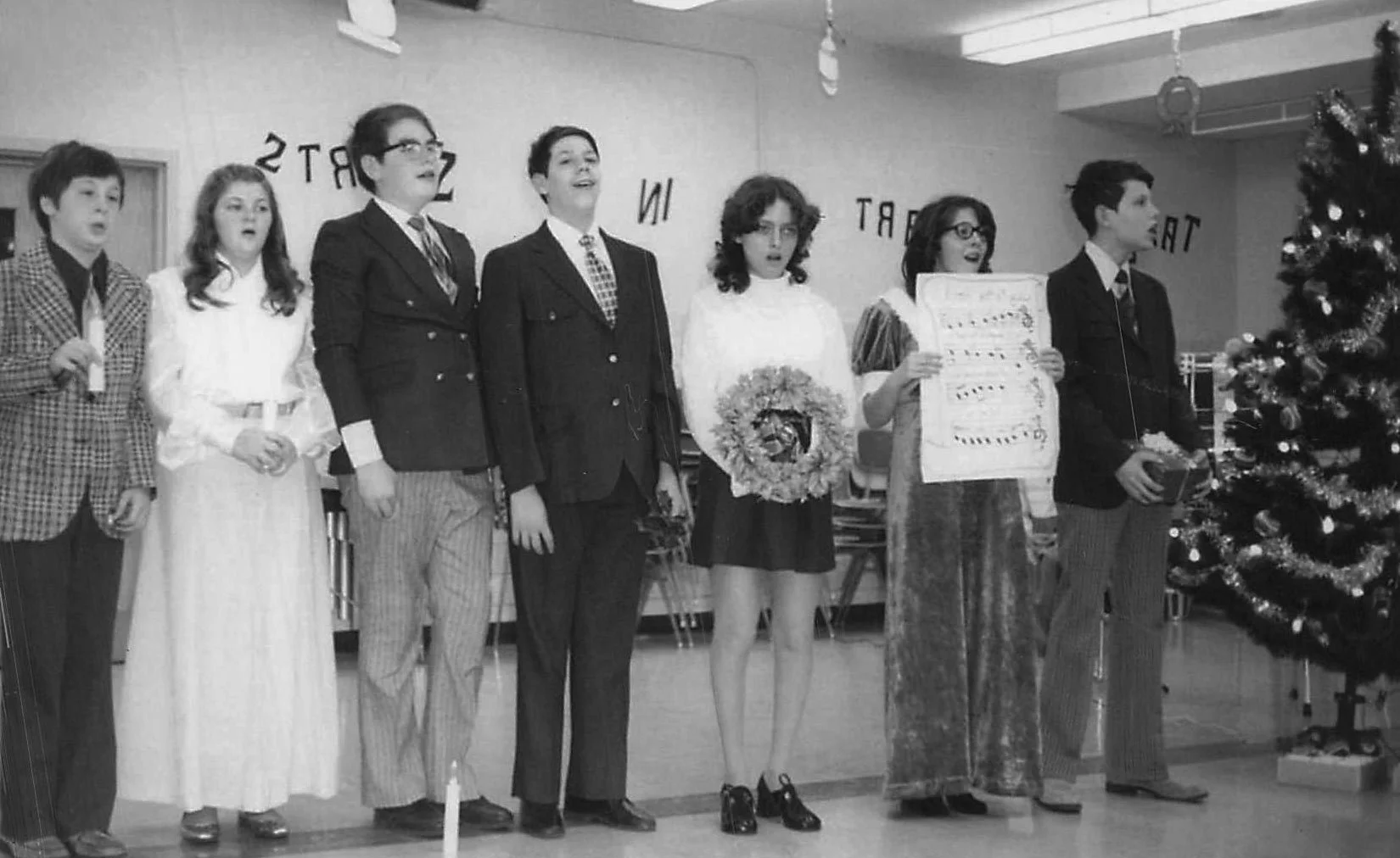 Black and white historical photo of seven children dressed in formal or festive clothes, standing in a line in front of a decorated Christmas tree, singing or performing at a holiday event in a classroom or community room.