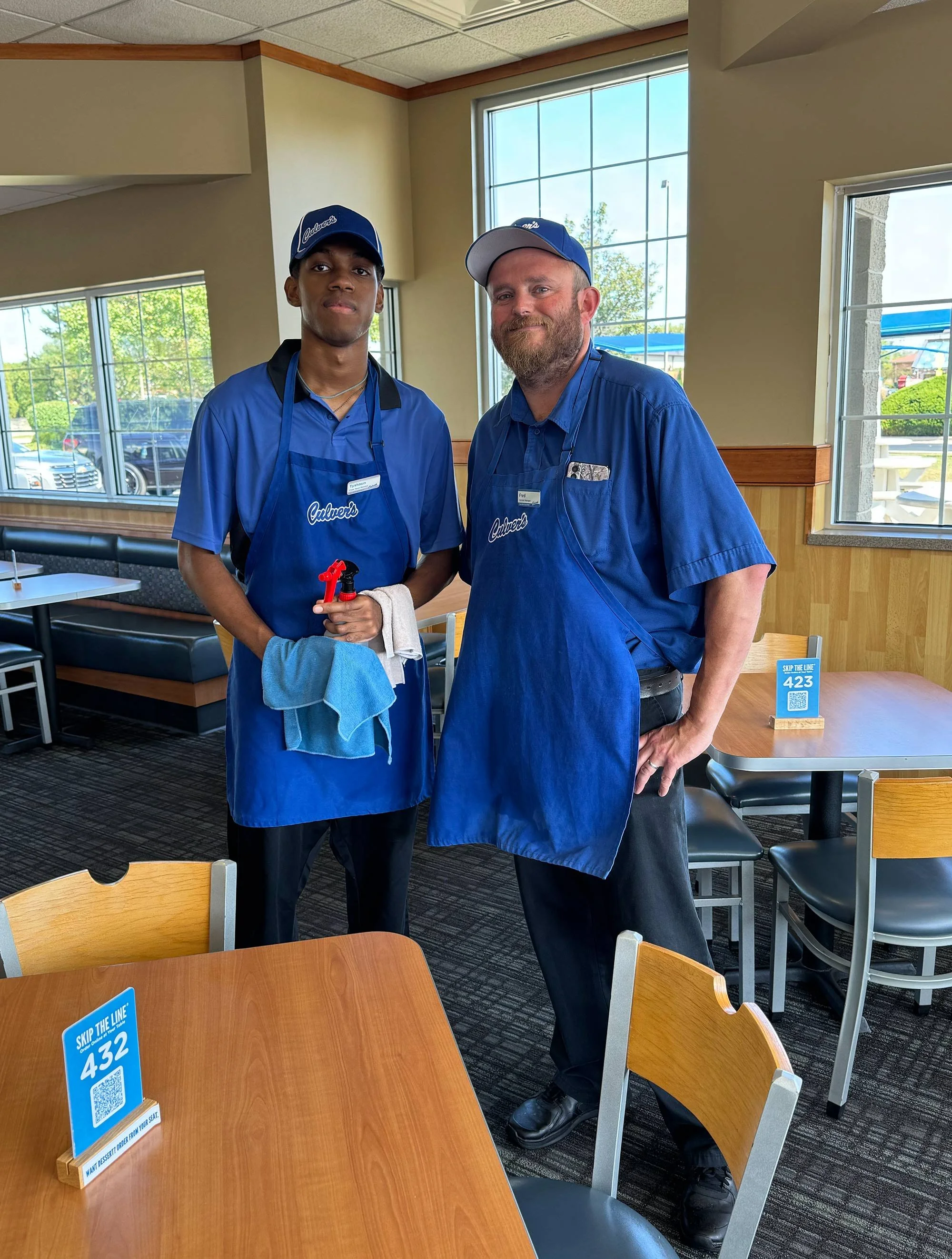 Two employees at a restaurant, one young and one older, both dressed in blue uniforms and caps, standing inside the restaurant with large windows and trees outside.