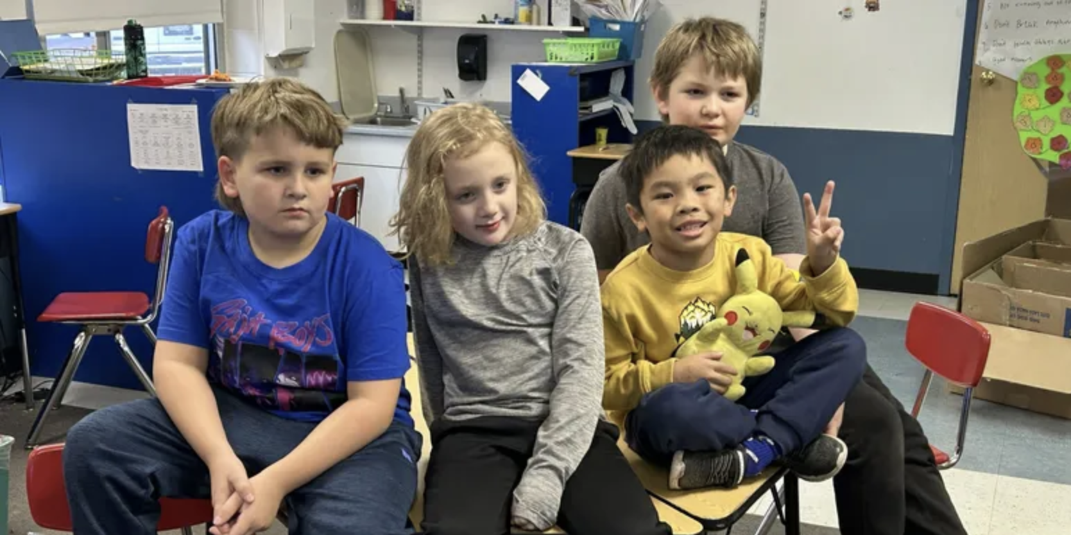 Five children sitting on a table in a classroom, with a girl in a gray shirt, a boy in a yellow shirt holding Pikachu plush, two boys behind them, and a boy in a blue shirt on the left. The classroom has toys, storage boxes, and a whiteboard.