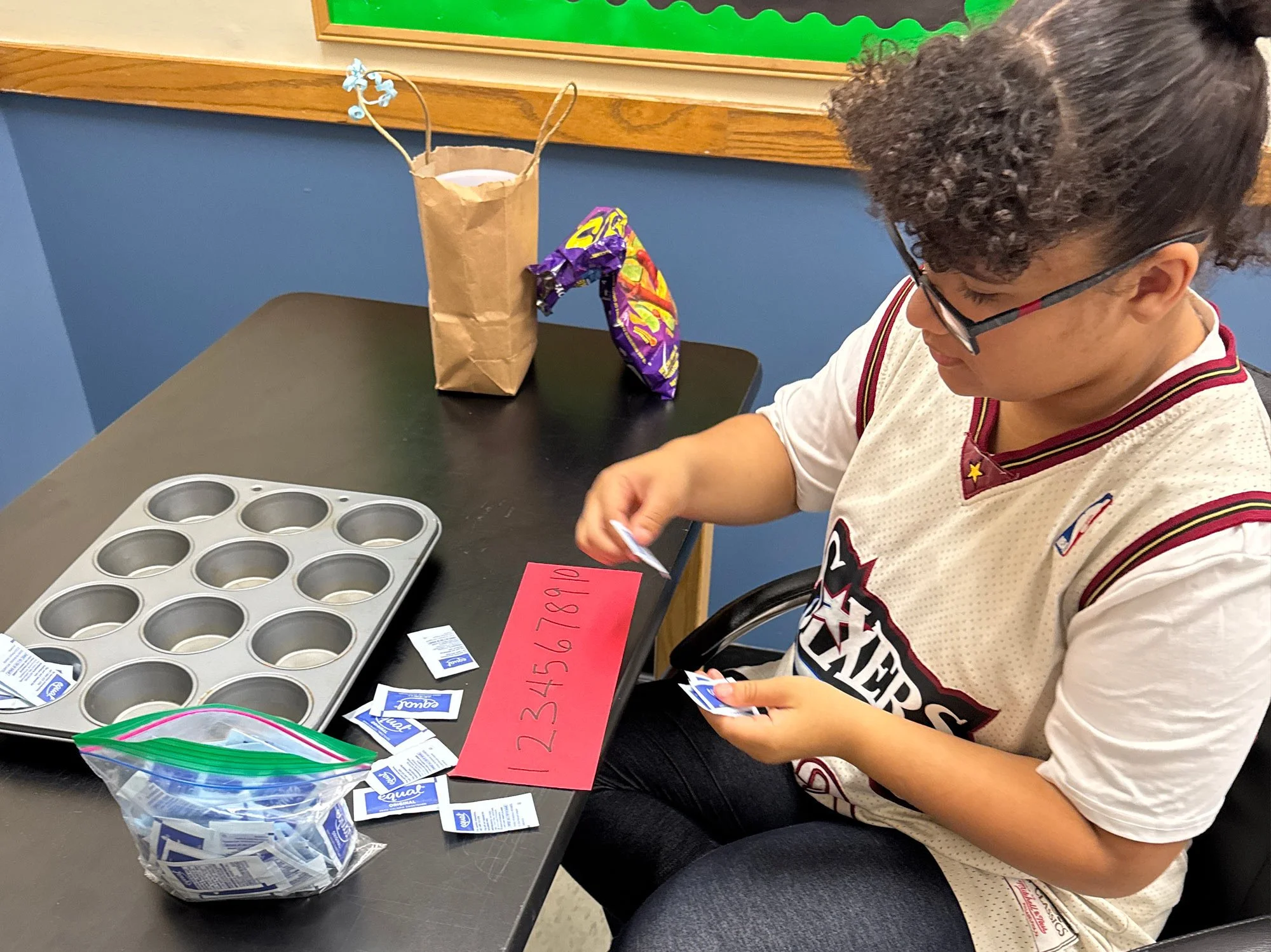 A young woman wearing glasses, a beige sports jersey, and dark curly hair sits at a desk with a muffin tin, packs of sugar, and a ziplock bag of sugar on the table. She is actively sorting the sugar packets.