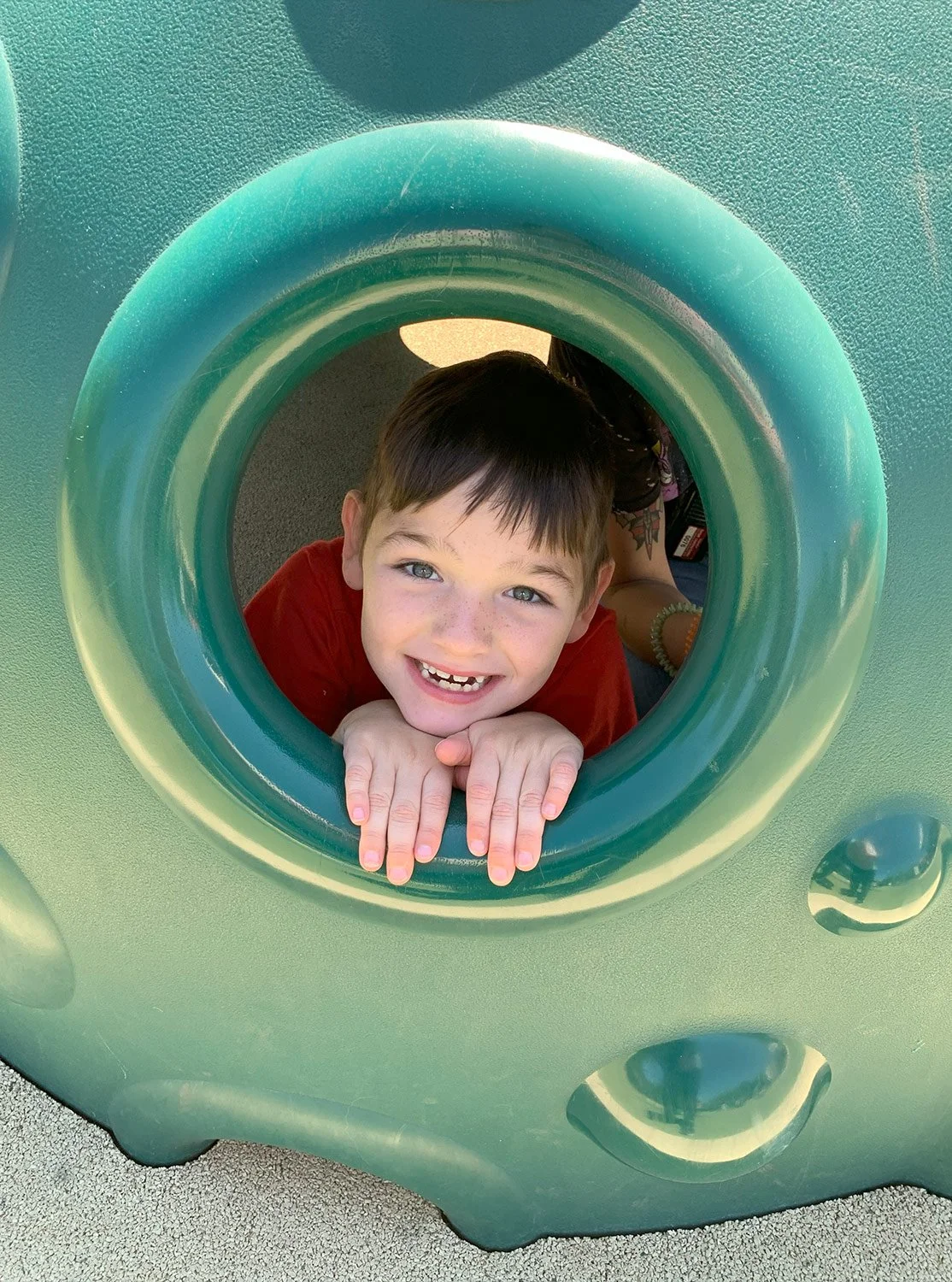 Child smiling and playing on playground, looking through a circular opening in a green structure.