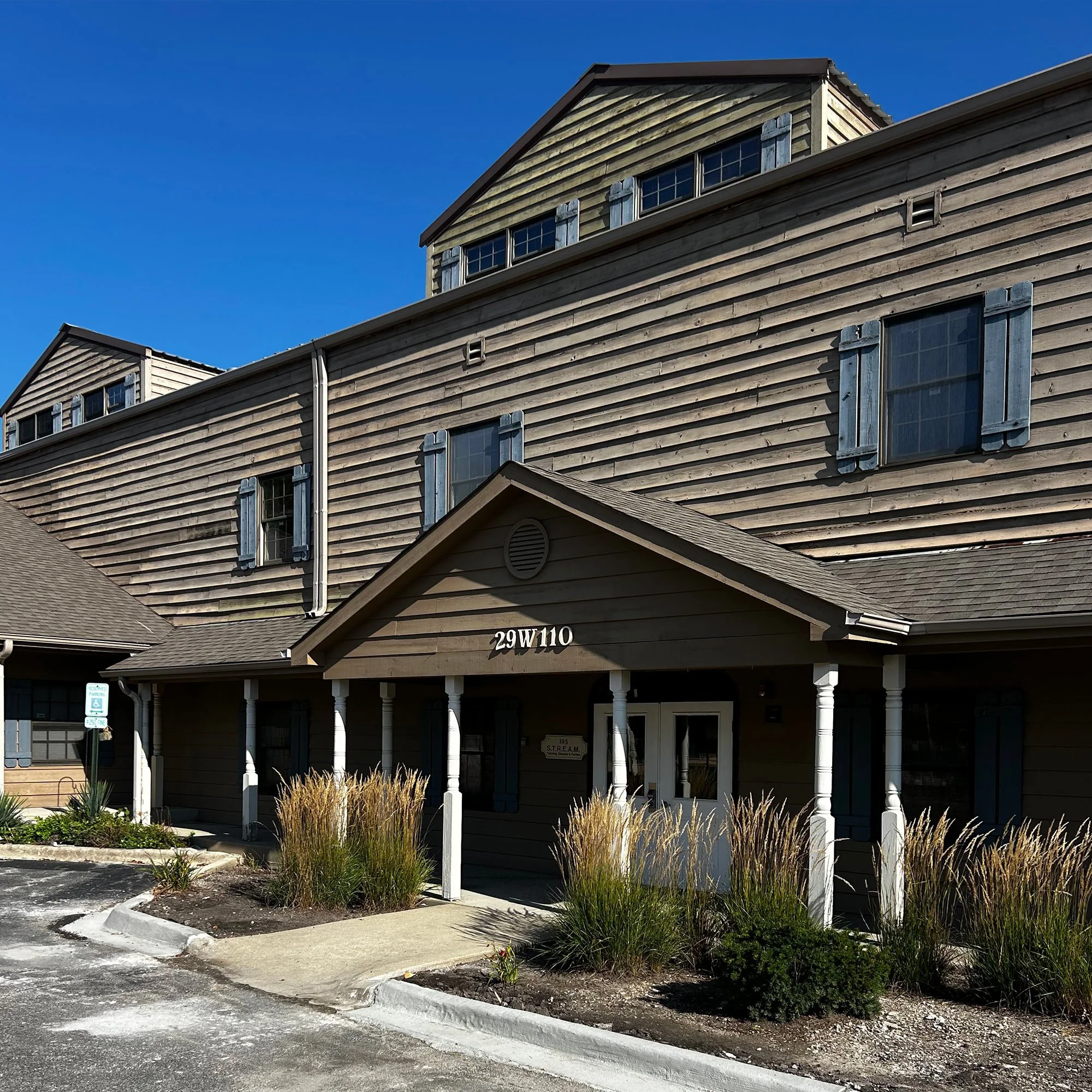 Image of a multi-story building with wooden siding, multiple windows with blue shutters, and a small porch with white pillars. There are parking signs and landscaped plants in front of the building under a clear blue sky.