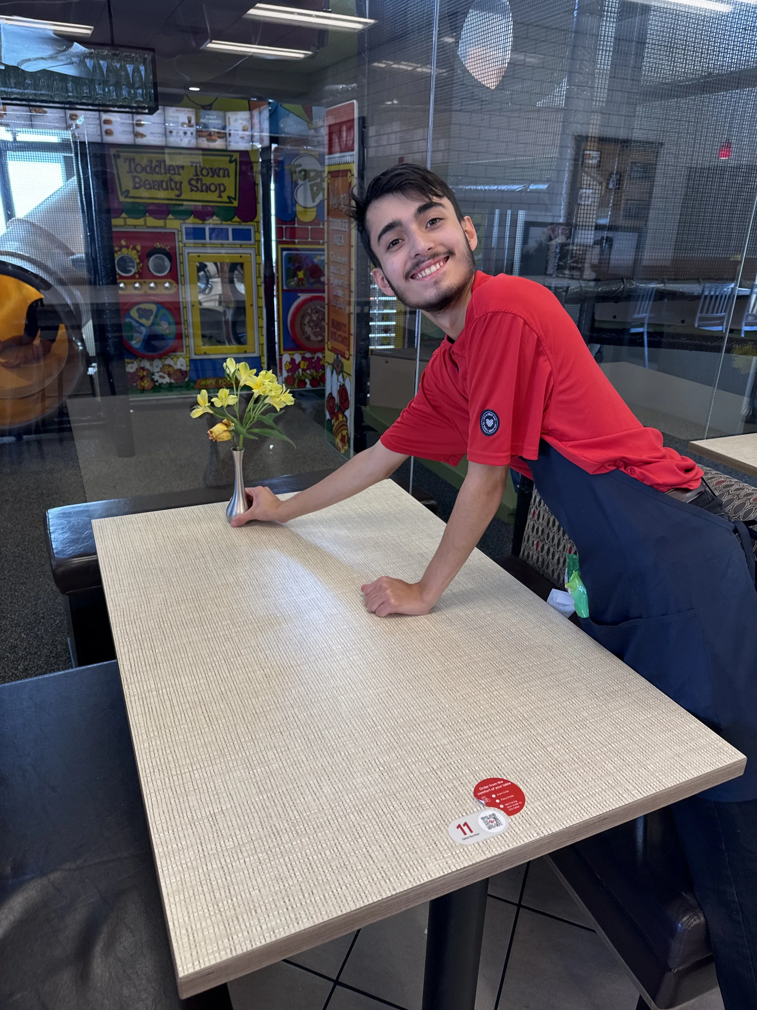 A young man in a red shirt leaning over a beige table with a small yellow flower arrangement in a silver vase in front of him. The background shows a colorful arcade game and a glass partition.