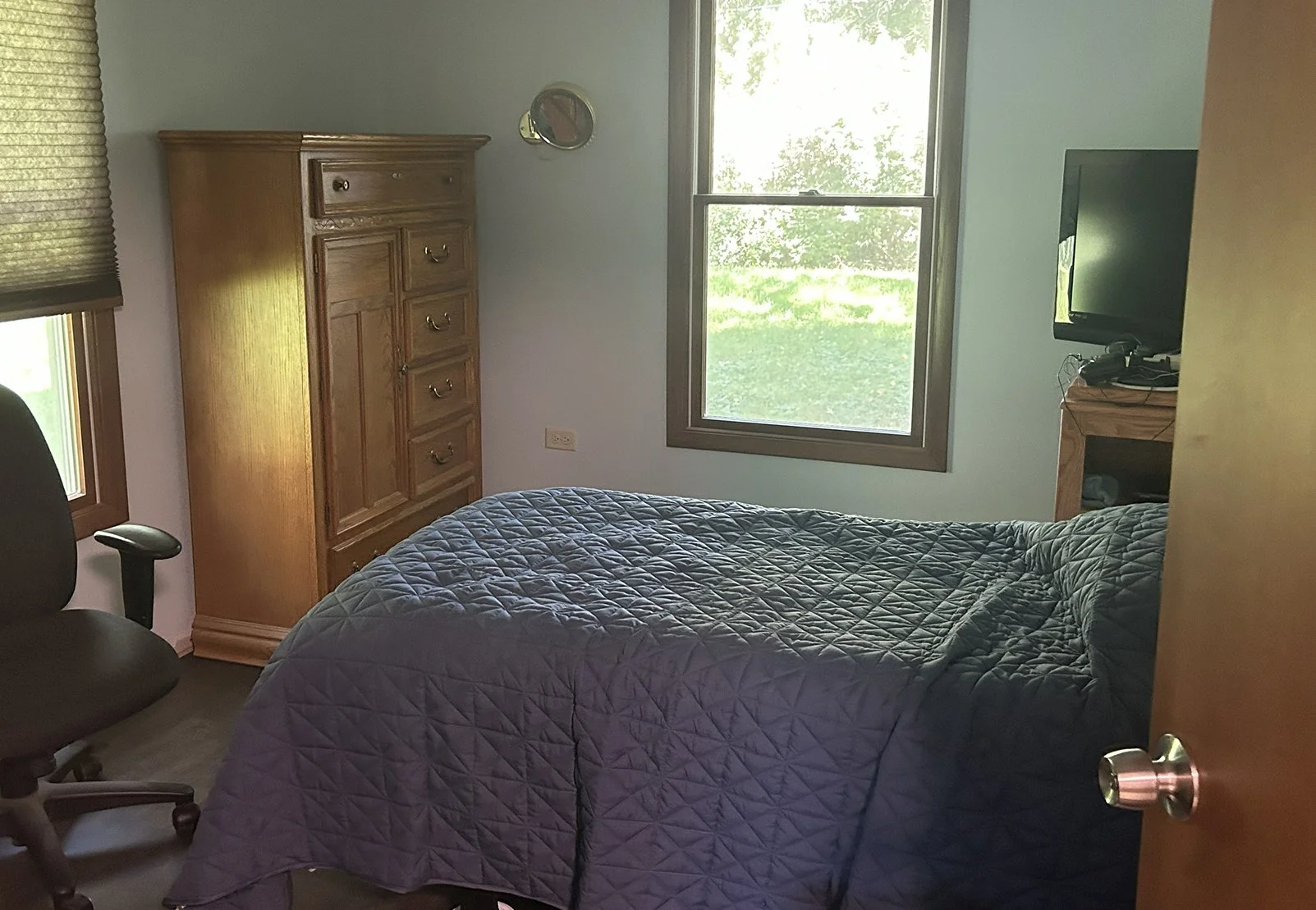 A bedroom with two windows, a wooden dresser, a bed with a dark quilt, a black office chair, and a wooden TV stand with a flat-screen TV.