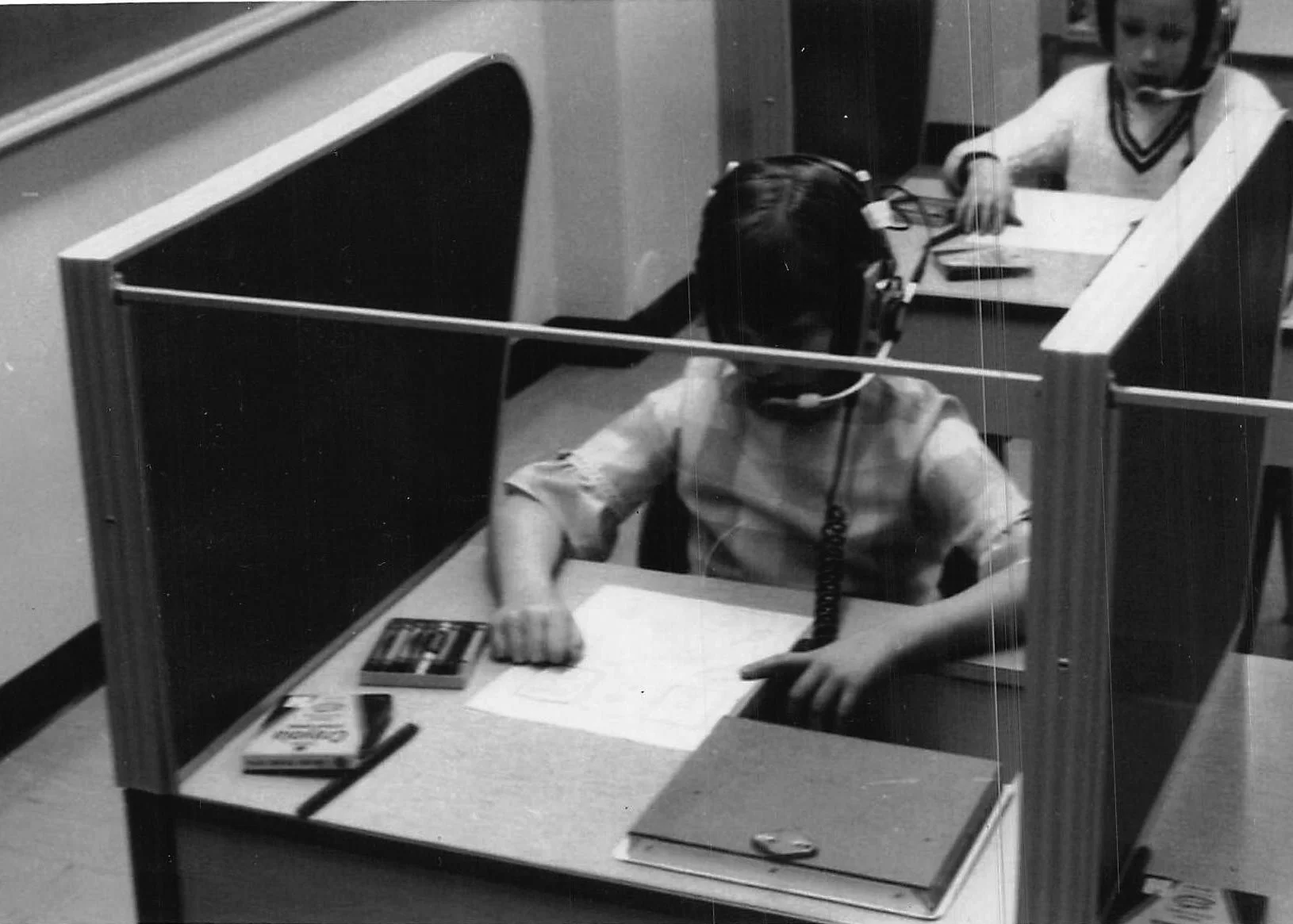 A young boy wearing a headset and speaking into a phone at a school desk in a cubicle, with another child in the background wearing headphones.