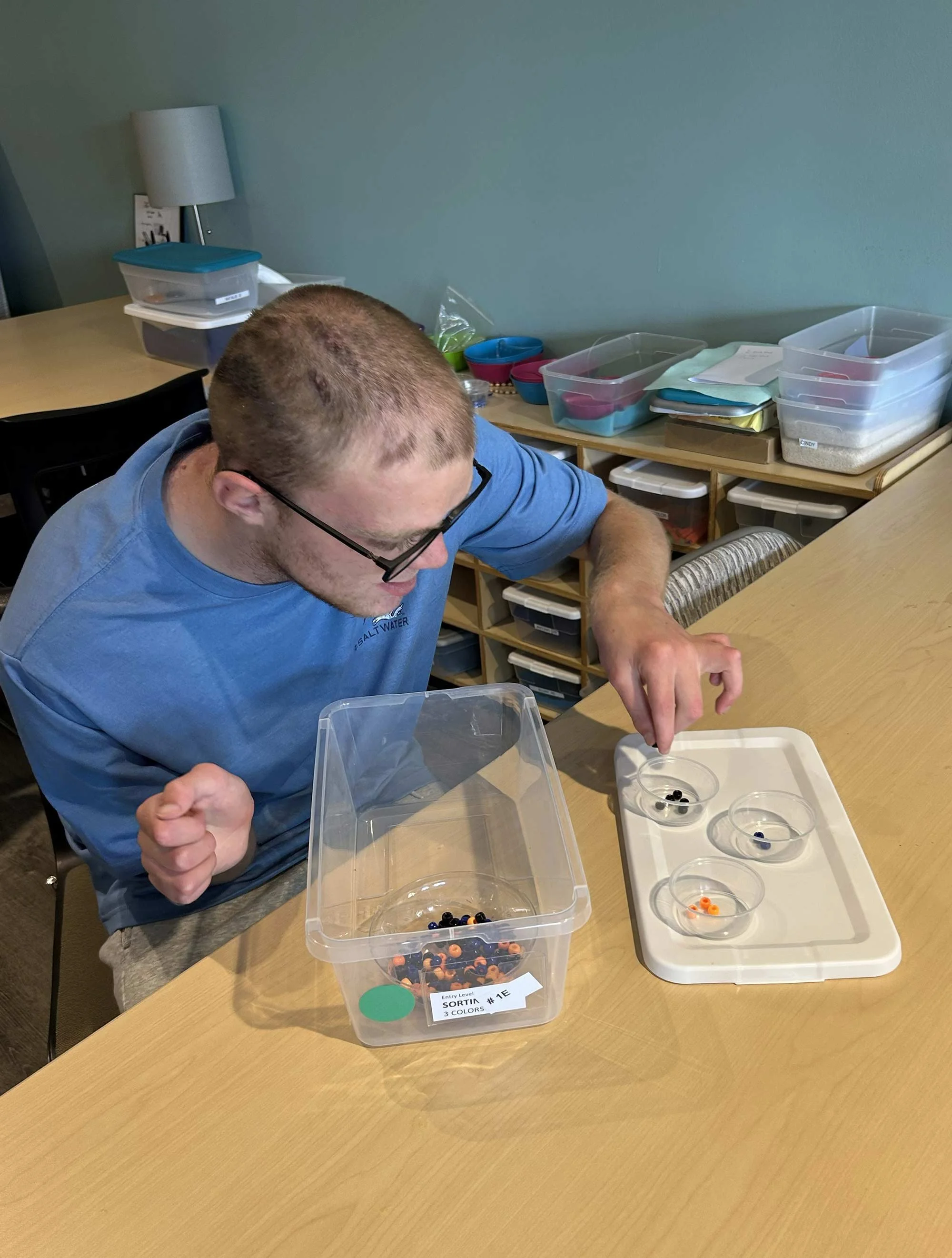 A man with glasses and a blue shirt sitting at a table, working with small beads in clear containers, with a collection of storage boxes and colorful bowls on the side.