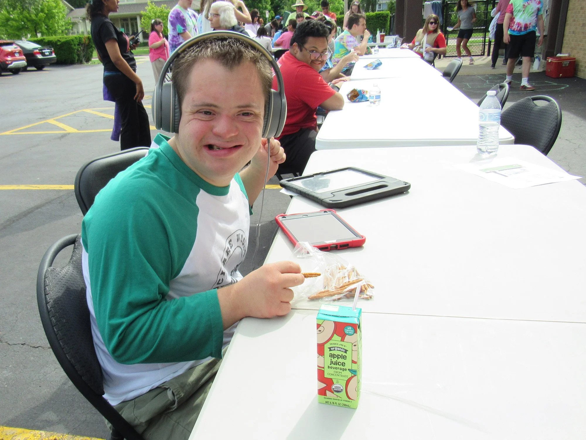 A young man with short hair, wearing a green and white shirt and large headphones, sitting at a long white table outdoors at a community event. He is smiling and holding a snack. On the table in front of him, there is a juice box, a black device, and a phone. Behind him, other people are seated at the table, eating, talking, and enjoying the event.