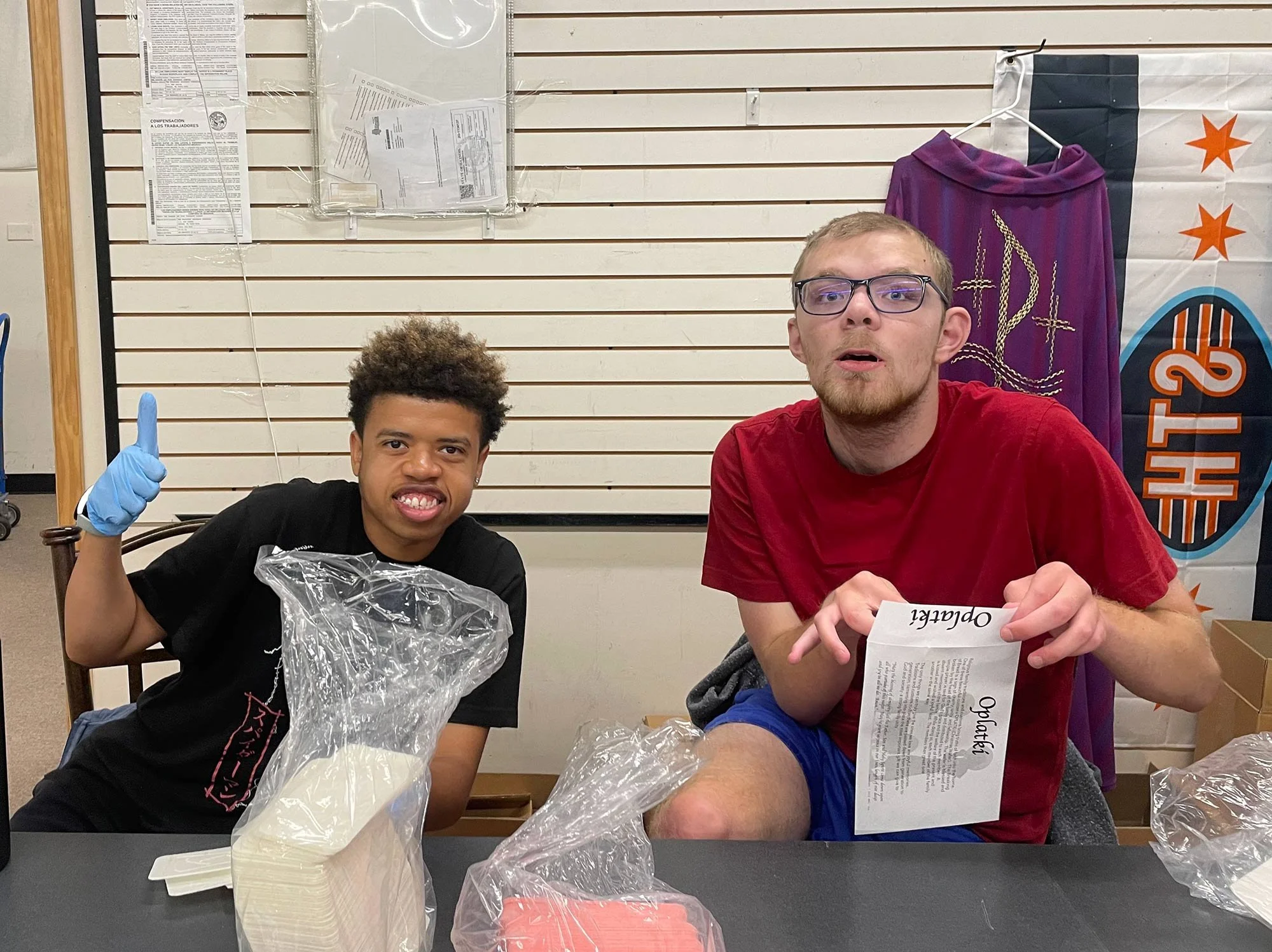 Two young men sitting at a table, sorting items. The man on the left is wearing a black shirt and a blue glove, smiling and giving a thumbs up. The man on the right has glasses, a red shirt, and is holding an envelope.