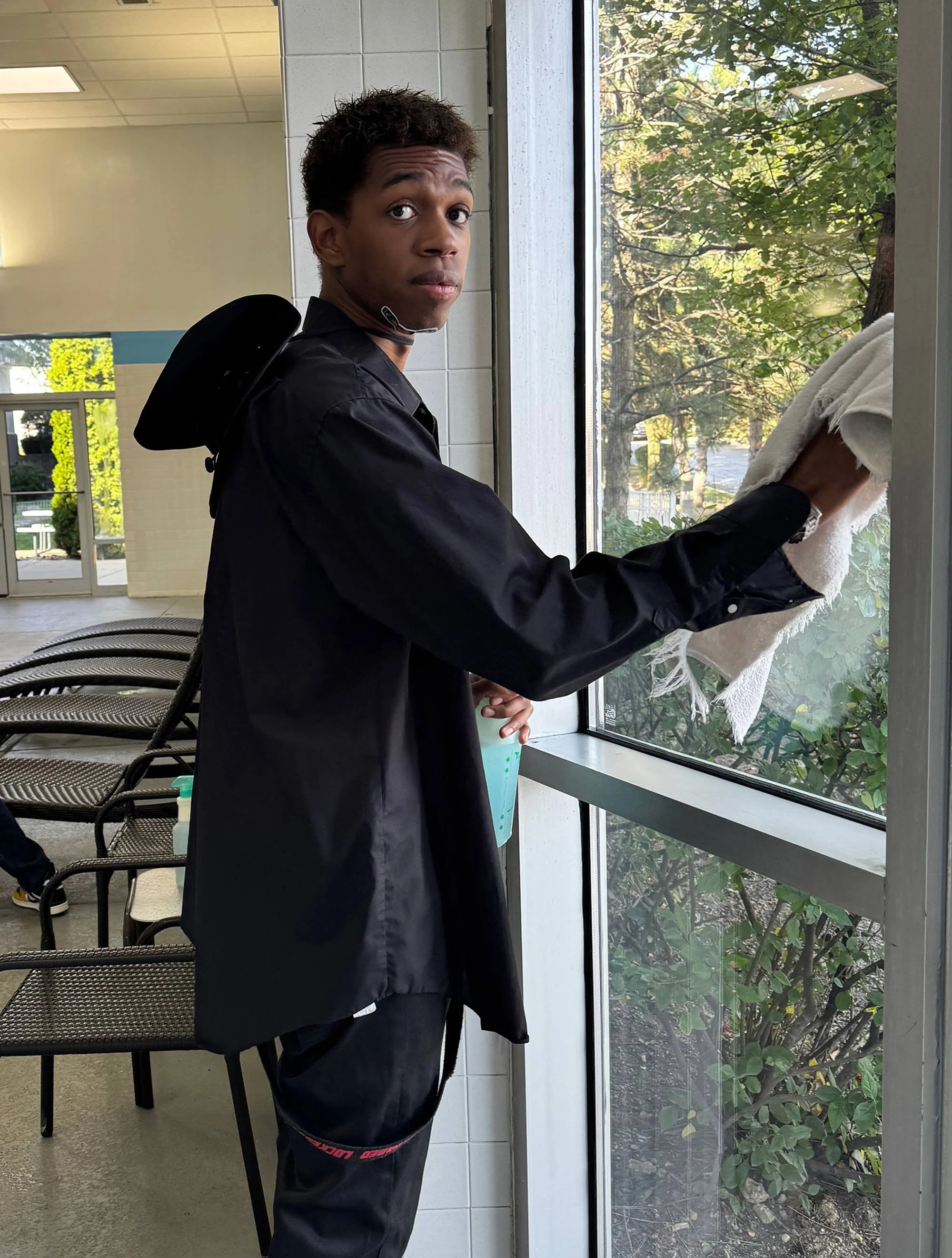 Young man with short dark hair, wearing a black jacket and dark pants, cleaning a large window with a white cloth inside a bright room with chairs, white tiled walls, and green foliage outside.