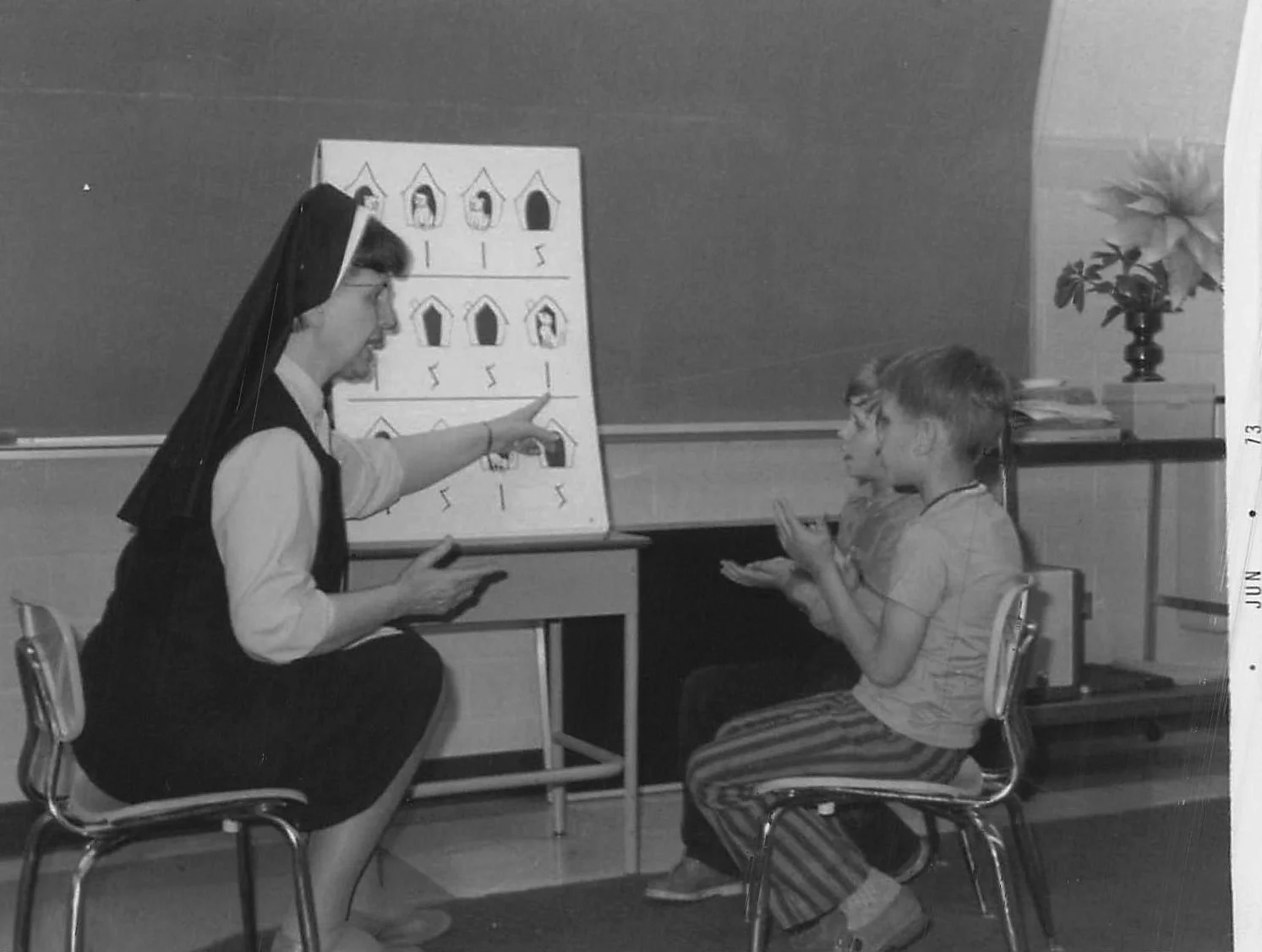 Black and white historical photo of a nun pointing at a board with diagrams of houses, explaining something to the children. The children are sitting on chairs, gesturing with their hands, and attentively listening.