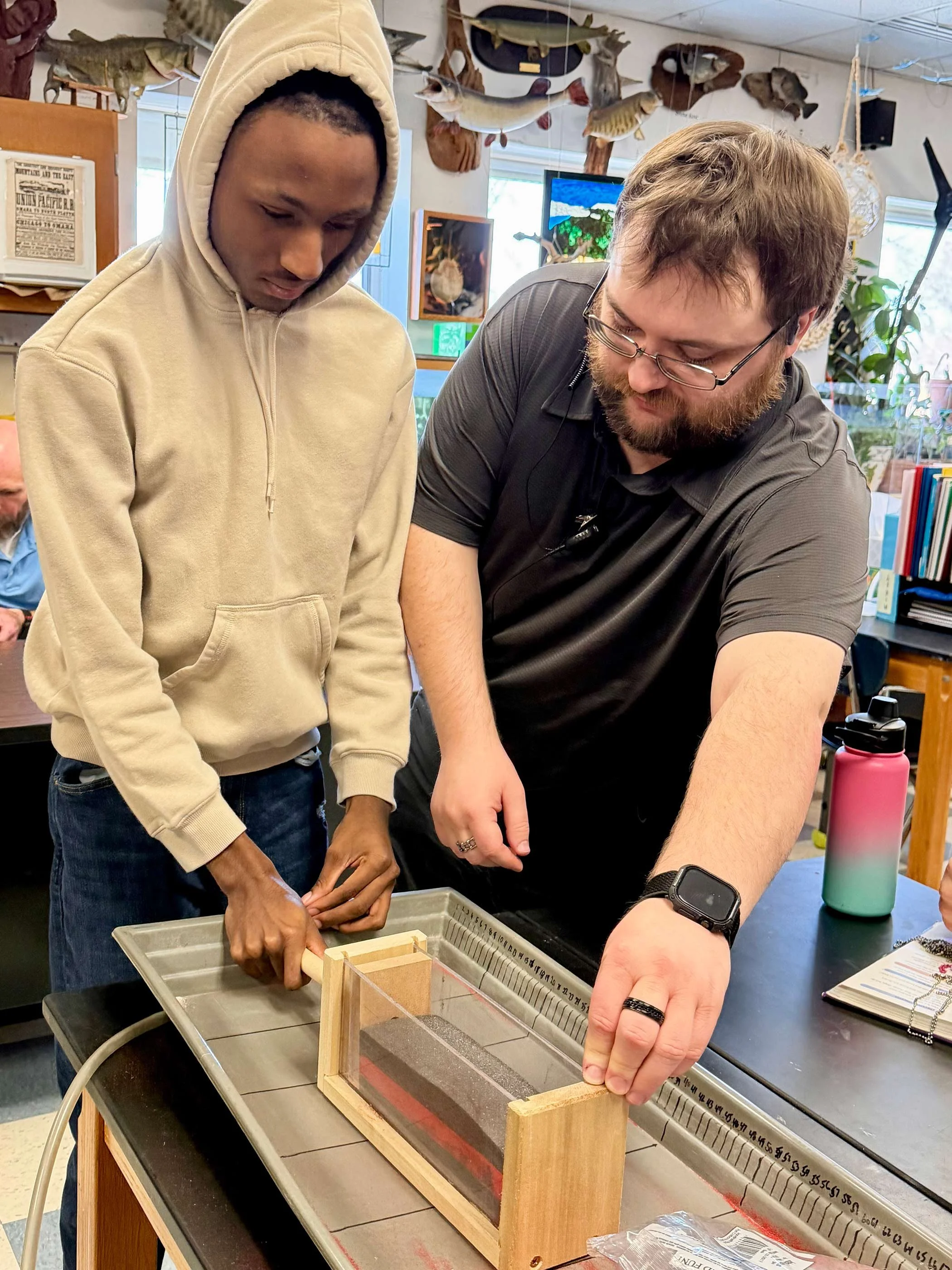 Two men are working together on a project at a table. The man on the left, wearing a beige hoodie, is holding a tool, while the man on the right, with glasses and a beard, is focused on a wooden frame with a clear panel.