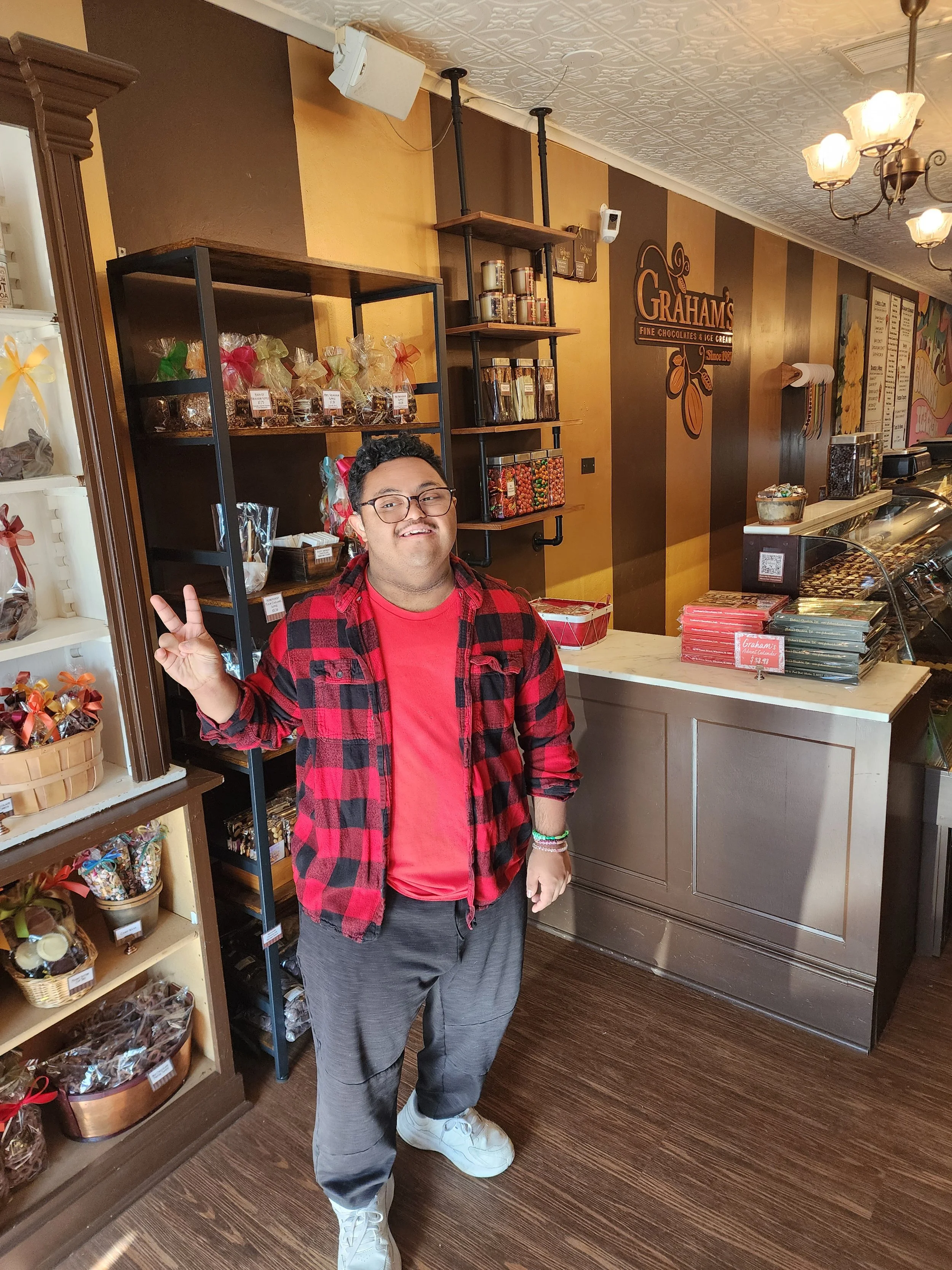A young man smiling and making a peace sign with his right hand inside a candy shop, standing in front of shelves filled with assorted chocolates and candies.